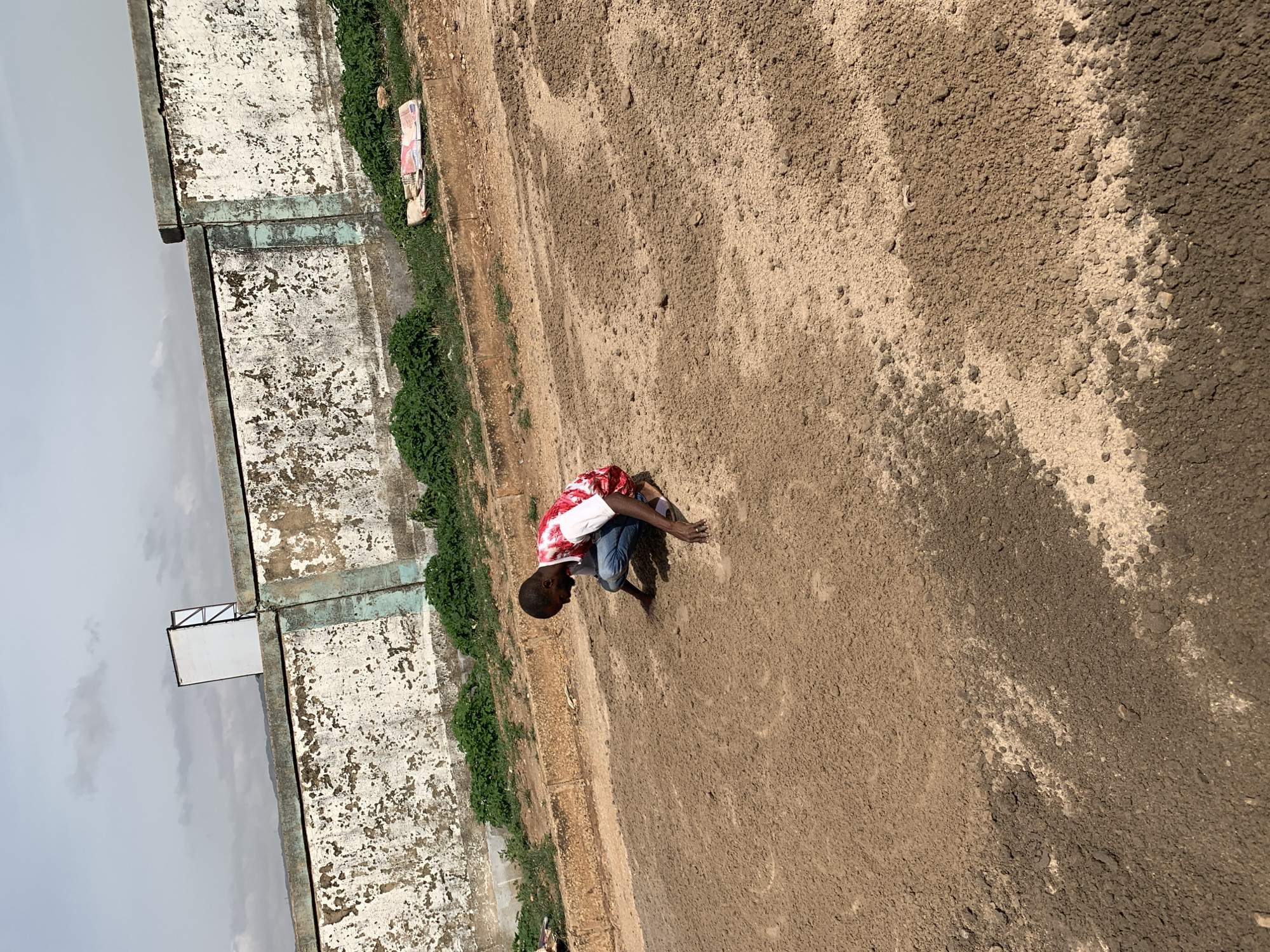 A young person crouches in a field of what appears to be dry, sandy soil. The individual is wearing a white and red patterned shirt and blue jeans. To the left of the subject is a deteriorated concrete wall covered in peeling paint with some vegetation growing along its base. To the far left, a light gray sky is visible. The location is Rafin Par, Nigeria.
