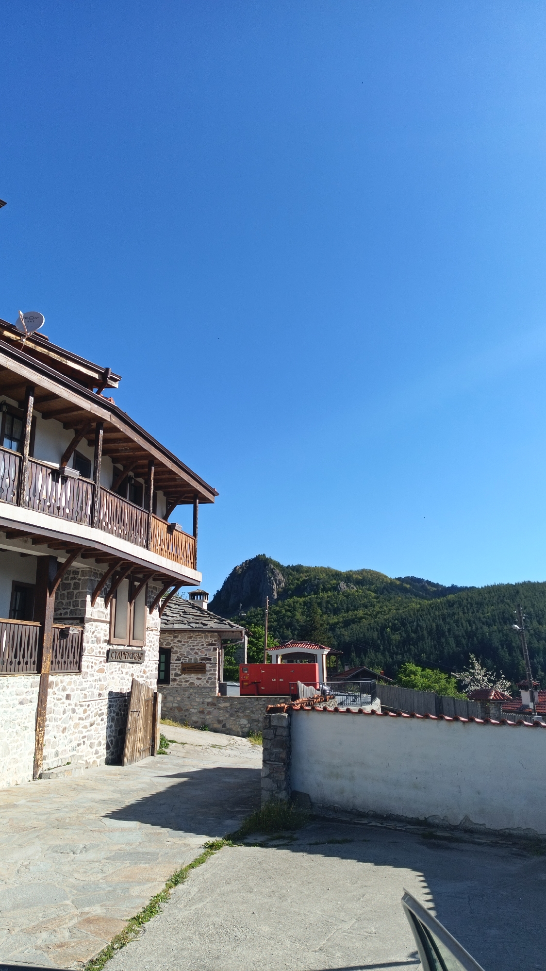 The scene is a street view in Vrata, Bulgaria. The sky is clear blue. On the left is a multi-story stone building with wooden balconies. Adjacent is a smaller stone building with a wooden door. In the background is a hill covered in green trees. A red object is behind the smaller building. A white wall runs along the right side of the street. The pavement is a mix of stone and concrete. The side mirror of a vehicle is visible in the bottom right.
