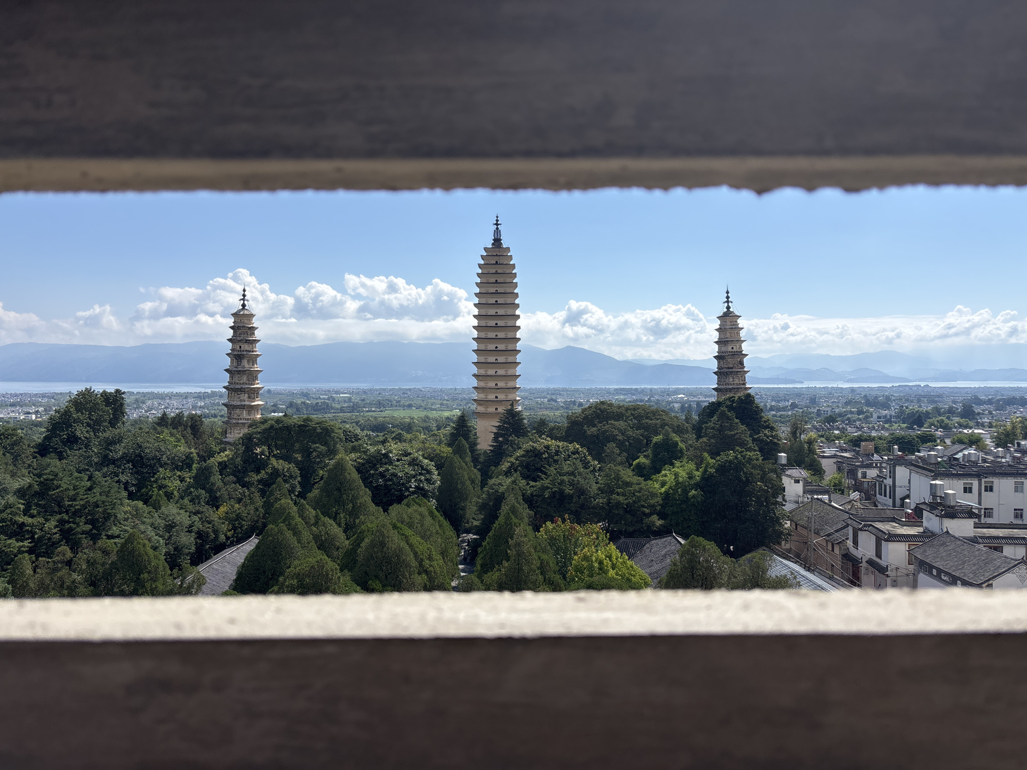 This image shows a scenic view of the Three Pagodas of Chongsheng Temple in Dali City, China, framed by what appears to be a balcony or window ledge. The three iconic pagodas, of varying heights and distinct architectural styles, stand prominently amidst lush green trees and foliage. In the distance, a city stretches out with traditional Chinese architecture featuring dark tiled roofs, and beyond that, a hazy mountainous landscape meets a clear blue sky dotted with fluffy white clouds. The scene evokes a sense of historical grandeur and natural beauty.