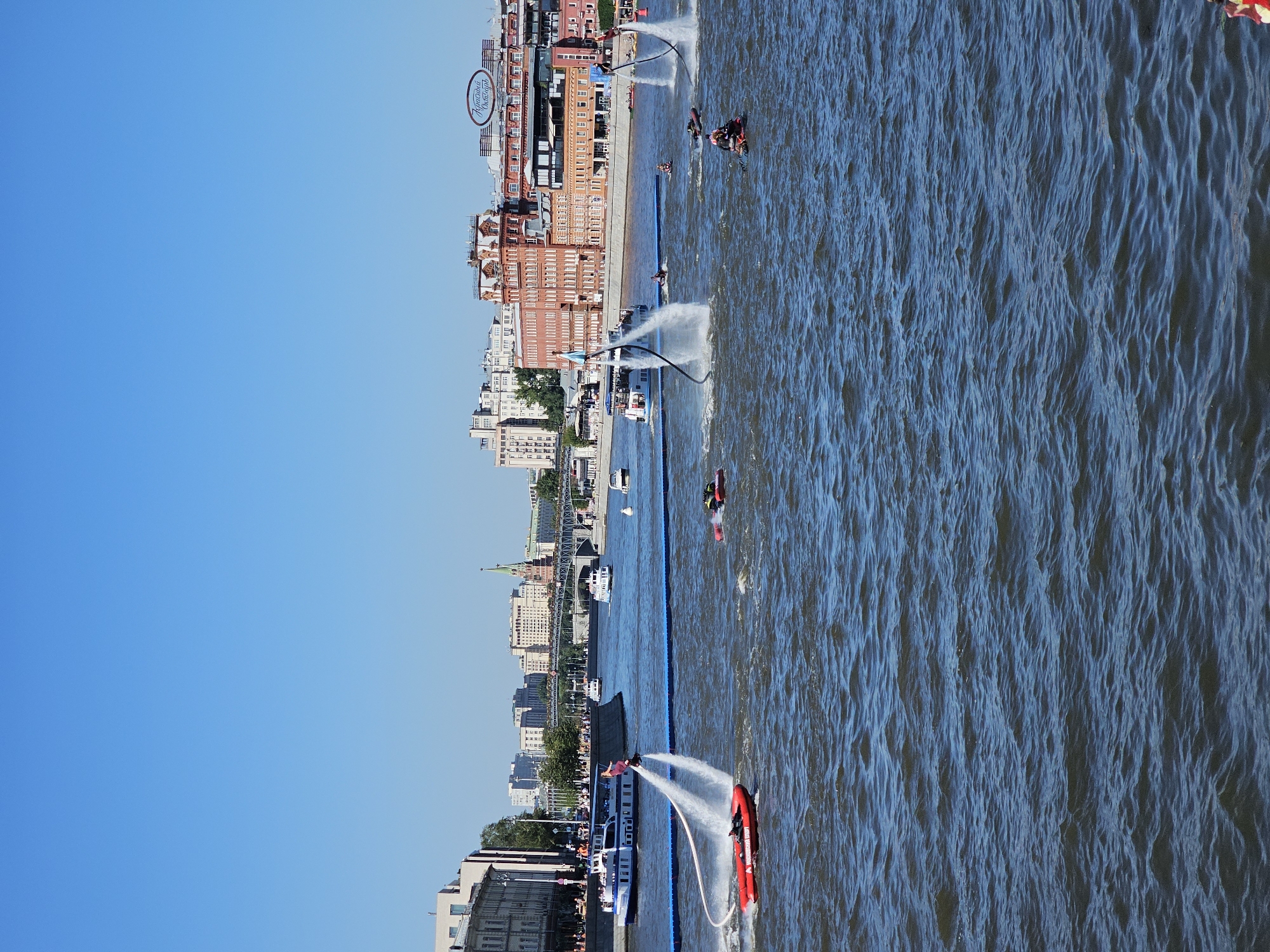 The image captures a sunny day on the Moskva River in Moscow, Russia, with several individuals engaged in water sports. In the foreground, a person on a jetpack is being propelled out of the water, creating a significant plume of spray. To the left, another person on a jetpack is also airborne, tethered to a red jet ski. In the background, a large white passenger boat is visible, navigating the river. The cityscape lining the riverbank includes various buildings, with prominent red brick structures and a bridge spanning the water. The sky is clear and blue, indicating good weather. Several other smaller watercraft, including what appear to be jet skis, are also present in the water, with some individuals riding them. The overall scene depicts a lively recreational activity on the river with the iconic Moscow skyline as a backdrop.