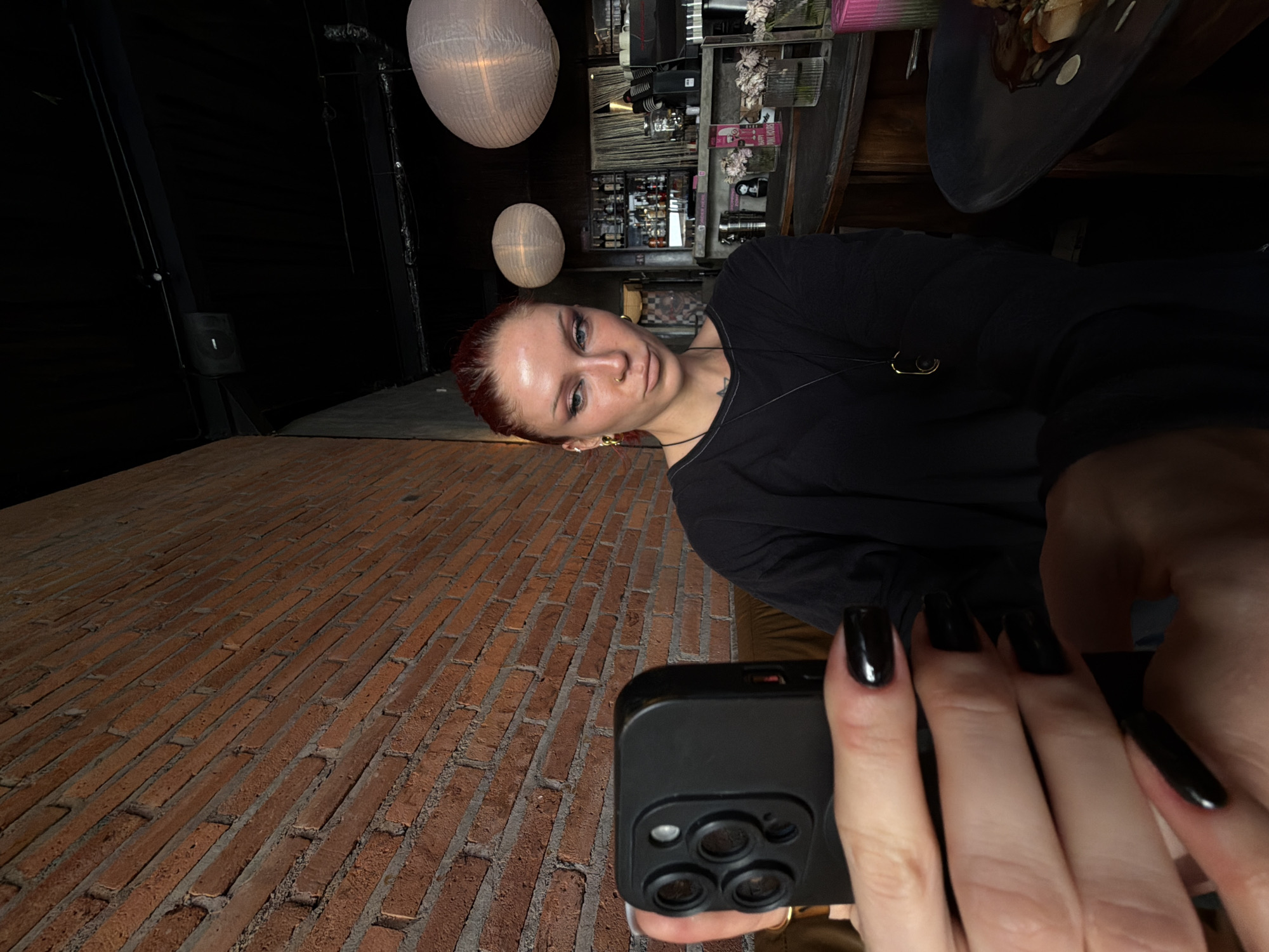 A woman with red hair pulled back and dark eye makeup is taking a selfie in a dimly lit cafe. She is wearing a black long-sleeved shirt and a necklace with a dark pendant. She has black painted fingernails. The cafe has exposed brick walls, dark ceilings with exposed beams and lights, and a bar area with shelves of bottles. Two large, round paper lanterns hang from the ceiling. In the foreground, her hand holds a black smartphone, showing the phone's camera lens. A dark plate with food is visible on a table to the right. The location is identified as North Kuta, Indonesia.