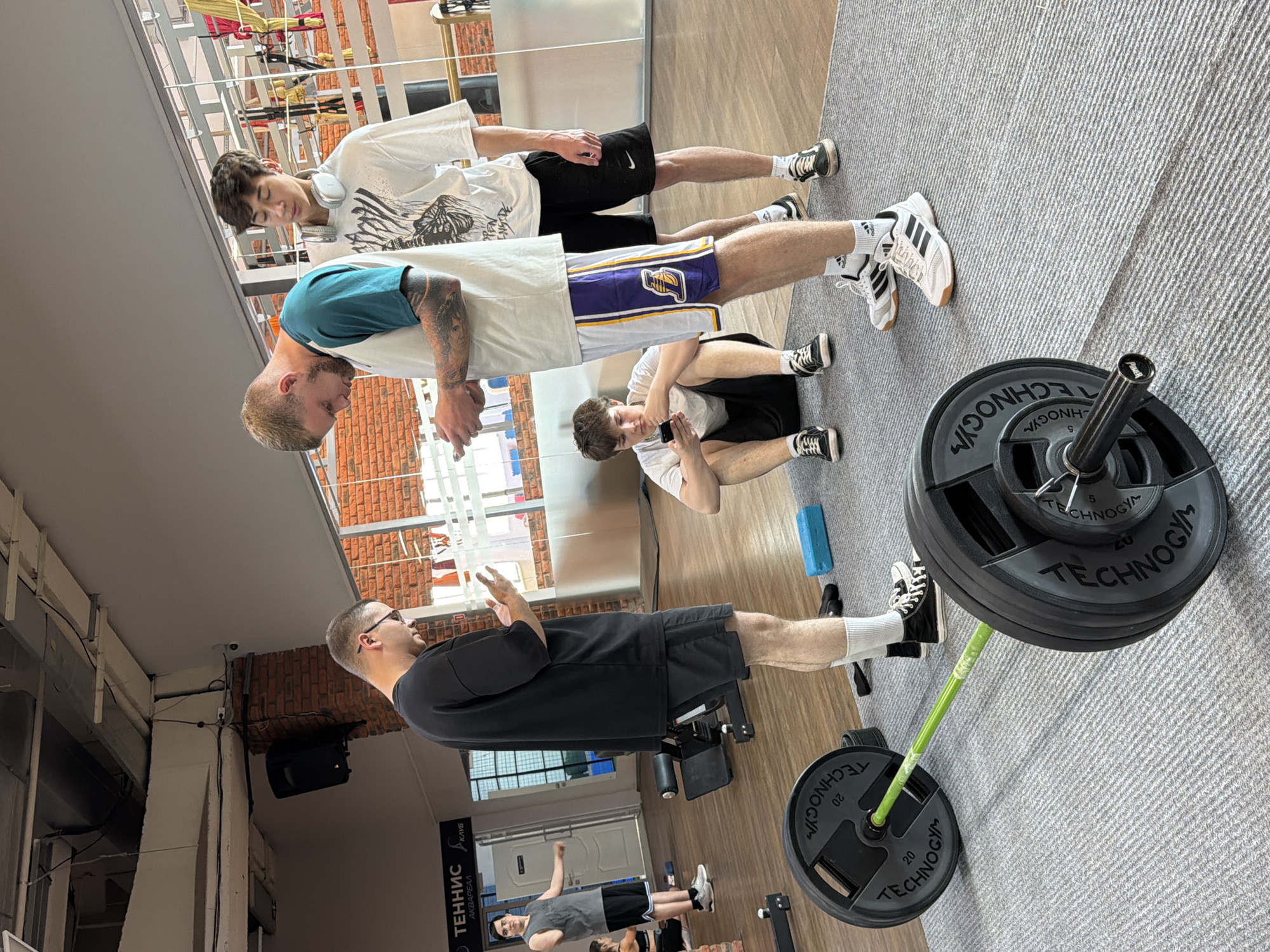 Here is a detailed description of the image:
**Location:** The image appears to have been taken in a gym, possibly in the city of Chaikovsky, Russia. This is inferred from the Cyrillic text on a sign visible in the background, which reads "ТЕННИС АКВАРЕАЛ" and the name "Чайковский" which means Chaikovsky.
**People:** There are four young men present in the image.
* The tallest man is standing and appears to be coaching or instructing the others. He is wearing glasses, a black t-shirt, and black shorts.
* Two young men are standing. One is wearing a white t-shirt with a graphic design, and black shorts and the other is wearing a blue t-shirt, white t-shirt, and Laker shorts.
* One young man is sitting down using his phone. He is wearing a white t-shirt and black shorts.
**Objects:**
* A barbell with weights on it is visible in the foreground. The weight plates are labeled "TECHNOGYM" and "20", presumably indicating the weight in kilograms.
* A blue foam roller is near the sitting person.
* There are various exercise machines and equipment visible in the background, as well as windows and some brickwork on the walls.
**Actions and Interactions:**
* The tallest man is gesturing with his hands, likely providing instruction or guidance to the other young men.
* The young man with the phone is looking down at it, likely taking a video of the group.
* The other two young men are listening.
**Environment:**
* The gym has a modern appearance with a gray floor. There are windows allowing natural light into the space.
**Overall Impression:** The image captures a scene of friends working out at a gym, with one acting as a coach or instructor. The environment is well-lit and equipped with modern exercise equipment.