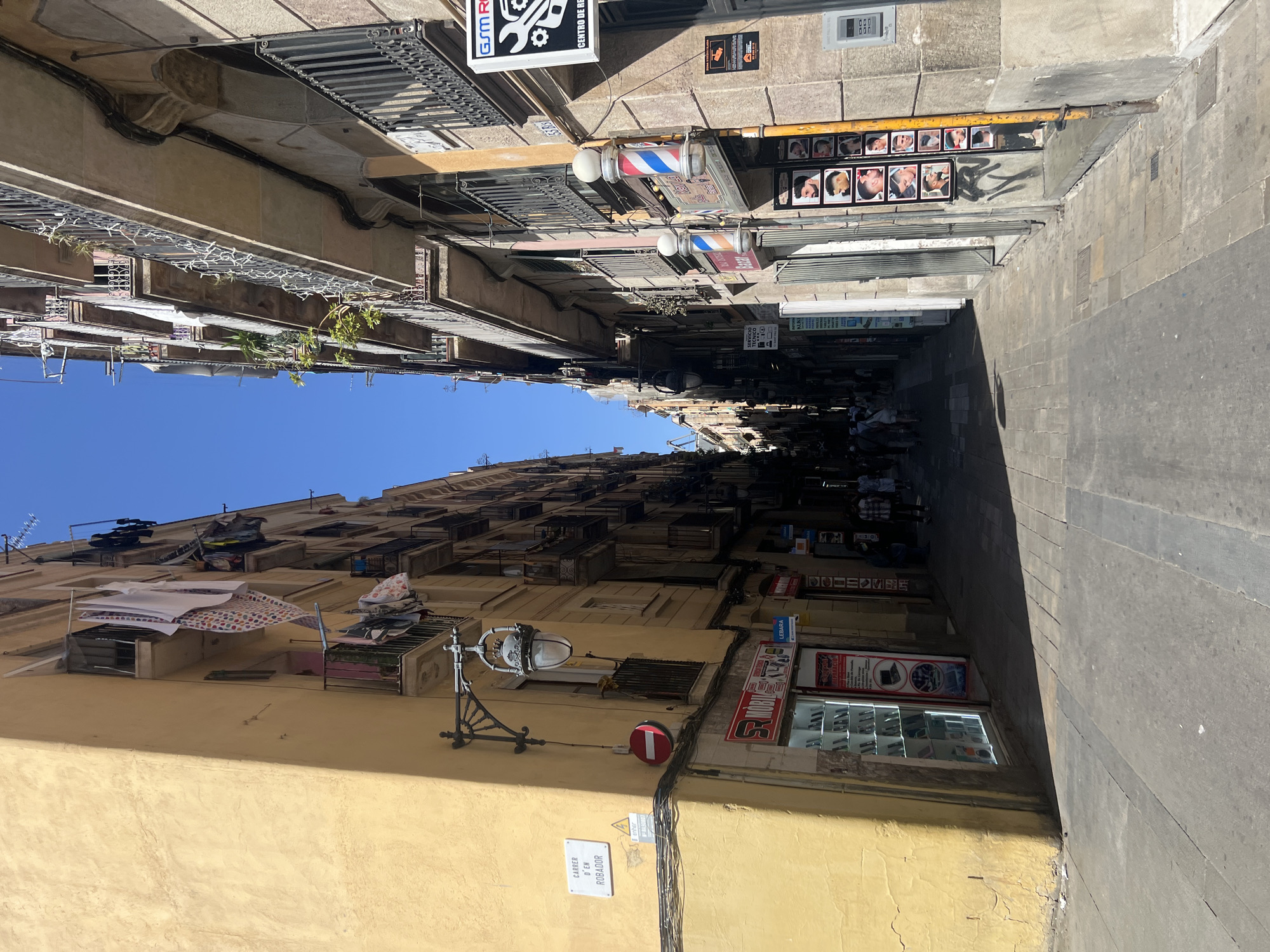 The image captures a narrow street in Barcelona, Spain, with tall buildings on either side. The building on the left is a vibrant yellow, while the building on the right has a more muted, stone facade. Laundry is visible hanging from balconies on both buildings, suggesting residential use. Several shops are present at street level, including a mobile phone store with a display window filled with devices and a barber shop featuring a traditional barber pole and a collage of men's hairstyles displayed on the wall. People are walking down the street, some appearing to be tourists or shoppers. The cobblestone pavement and the architecture are characteristic of older European cities. The sun is casting strong shadows, indicating it is daytime.
