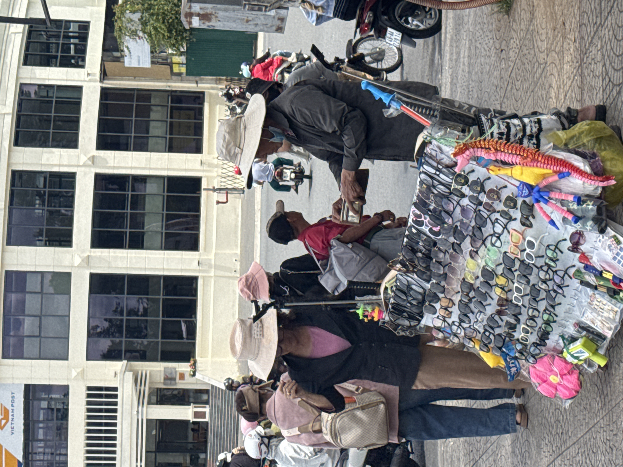 The image captures a street scene in Da Lat, Vietnam, likely near a commercial area given the architecture of the building in the background. A sidewalk vendor is the central focus, pushing a cart filled with an array of sunglasses and other small merchandise. The vendor, a man wearing a wide-brimmed hat and a dark long-sleeved shirt, is interacting with a customer who is partially visible and wearing a red shirt.
Several other individuals are present in the scene, most of them on motorcycles, indicating typical street traffic. One woman, wearing a large, light-colored hat and a dark jacket over a pink shirt, is standing near the sunglasses cart, appearing to be a potential customer or bystander. Another woman in a pink hat is visible behind her. A younger person in a red shirt and a cap is also in the vicinity of the vendor.
The environment is an urban setting with a modern building featuring large, dark-tinted windows. The sidewalk has a patterned tile surface. The overall impression is of a bustling street with pedestrian and vehicular activity, characteristic of a Vietnamese city. The cart is decorated with brightly colored items, including stuffed toys and plastic accessories, alongside the sunglasses.