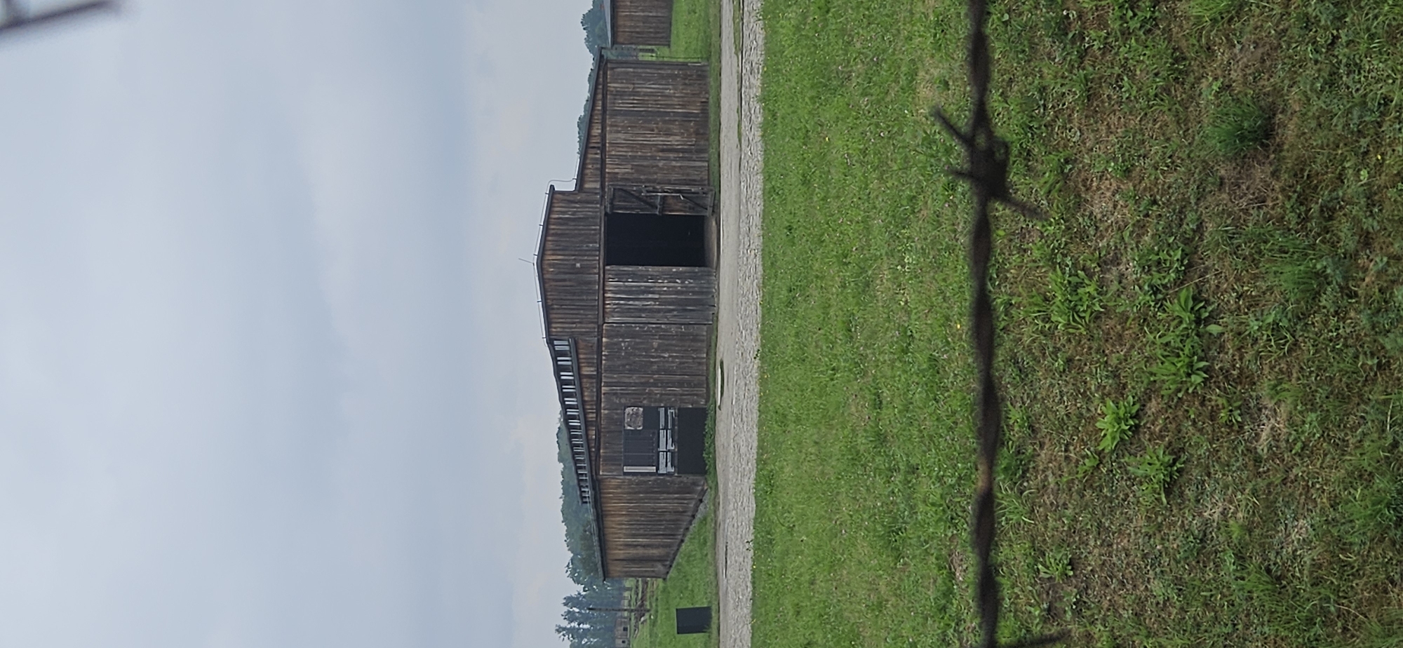 The image depicts a long, wooden barrack building with a central opening and two side doors, located in Brzezinka, Poland. The building has a pitched roof with a row of windows running along the ridge. The walls are constructed of vertical wooden planks. In front of the building, there is a paved area. To the left of the barrack, a black rectangular object is visible. The foreground is dominated by green grass with some patches of dry earth, separated by a barbed wire fence, the wire of which is blurred and out of focus. In the background, more trees and the tops of other wooden structures can be seen, along with a cloudy sky.