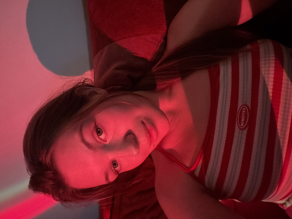 A woman lies on her side, facing the camera. She has light skin, brown hair, and green eyes. She wears a red and white striped tank top with a "nineties" patch. Red light bathes the scene, casting a pinkish hue on her face and body. Part of a red couch or pillow is visible behind her. A blue light shines behind her head.