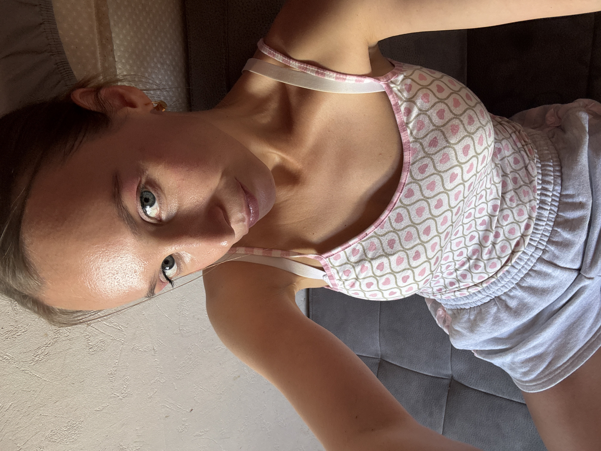 A young woman is sitting on a grey upholstered couch, looking towards the camera. She has blonde hair pulled back, blue eyes, and is wearing a white tank top with a pattern of pink hearts and light brown lines, and light grey shorts. She has a small gold hoop earring in her right ear. The background consists of a textured white wall on the left and the couch she is sitting on and some indistinct patterned fabric on the right. The lighting suggests it is daytime. There is no indication of a specific location within Überherrn, Germany.