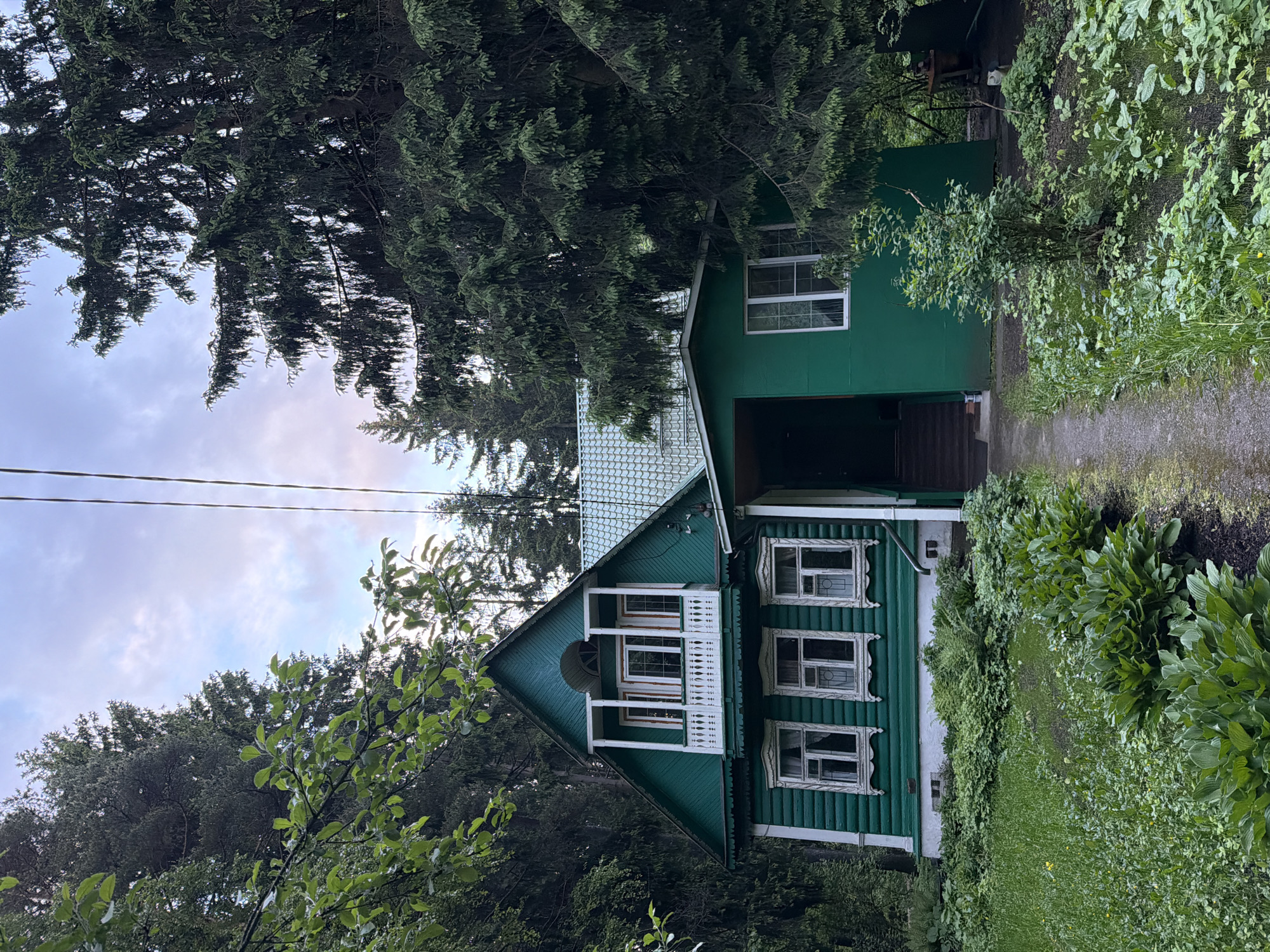 A green, two-story house with white trim is nestled amongst trees and foliage in Trudovaya, Russia. The house features multiple windows, a small balcony on the second floor, and a garage or covered area on the right side. A dirt path or driveway leads towards the house. Overhanging trees partially obscure the house and sky. Electrical lines are also visible.