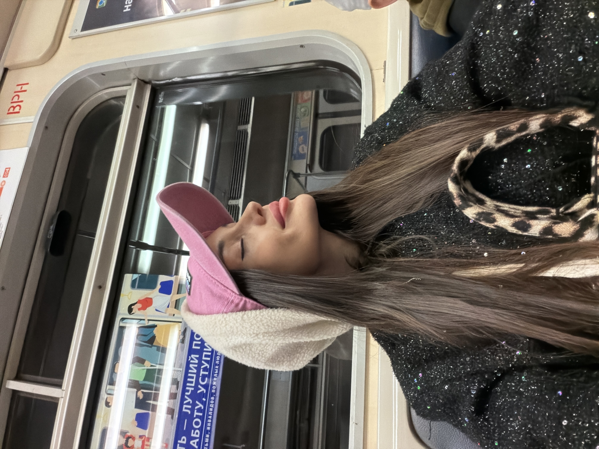 A woman is sitting on a Saint Petersburg, Russia metro car. She is wearing a pink hat with a white fur lining and a black sweater. The metro car has signs in Russian on the walls. There are multiple windows on the train car with handles.