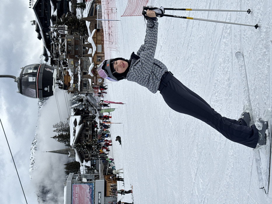 The image depicts a winter scene on a ski slope in Courchevel, France. In the foreground, a person in ski attire is positioned on skis, leaning to the left with ski poles held in their right hand. The individual wears a grey and white patterned ski jacket, dark blue ski pants, ski boots, a purple and grey ski helmet with goggles resting on top, and a dark head covering beneath the helmet. Their head is turned towards the viewer, with a slight smile and tongue partially extended.
The background shows a bustling ski area with multiple individuals skiing, standing, or sitting on the snow. To the left, a large signboard displays "CANYON DE YEPA" and "ALPIN" text. Further back, numerous traditional alpine chalet-style buildings with snow-covered roofs are visible. A prominent dark grey gondola cabin, marked with "COURCHEVEL", is suspended by a cable overhead, moving from left to right across the frame. A church spire is visible among the buildings on the left side of the slope. On the right, signs for "LANA" and "LE DENALI" (identified as luxury hotels in Courchevel 1850) are visible on multi-story buildings. Distant mountains, partially obscured by low-hanging clouds, form the horizon under an overcast sky. The ground is uniformly covered in packed snow, with faint ski tracks visible. Red safety netting is present on the far right.
