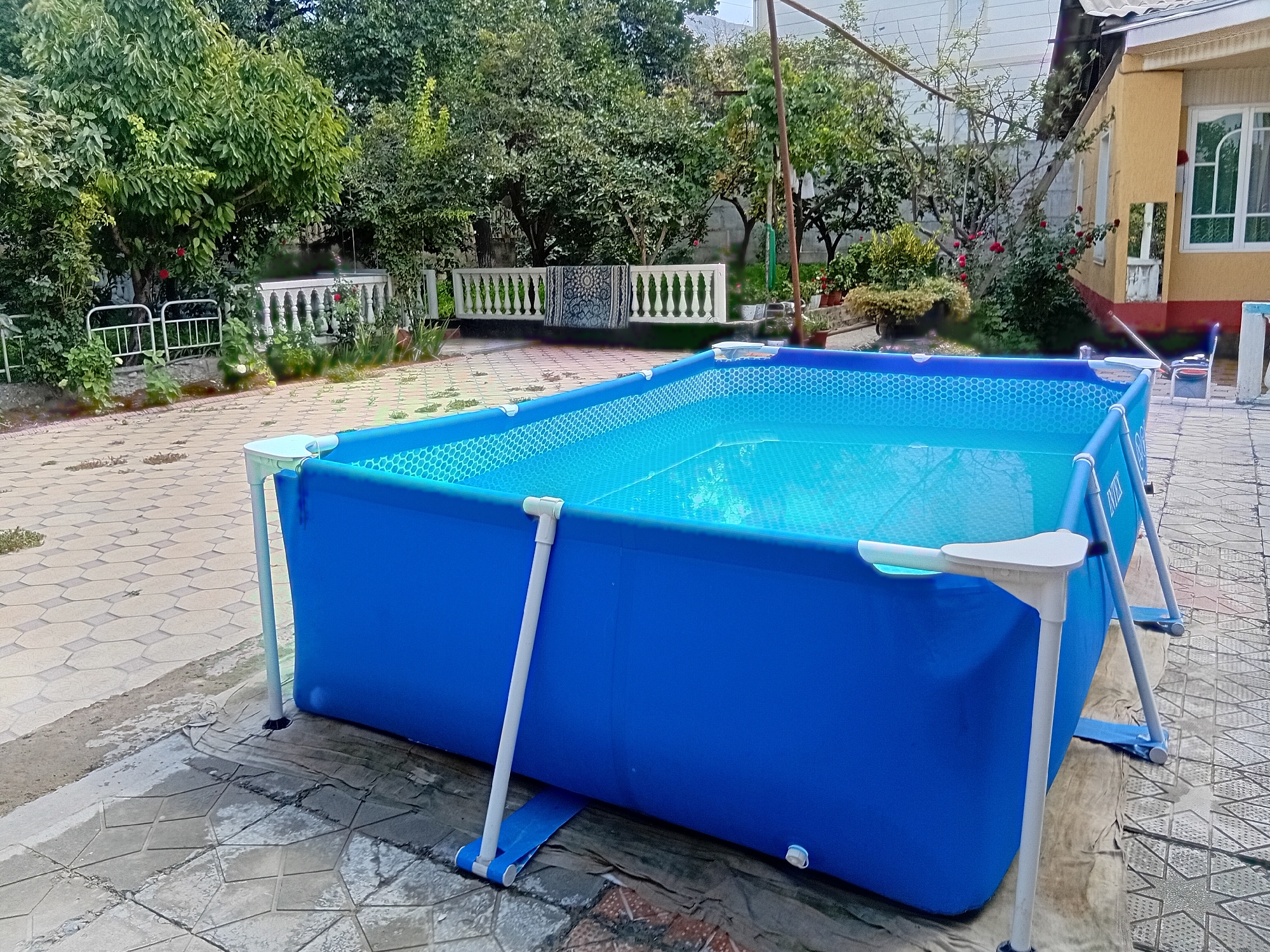 The image shows a large, rectangular, above-ground blue swimming pool filled with water in a backyard setting. The pool has white supporting poles and is situated on a paved patio area. In the background, there is lush green foliage from trees and bushes, a white decorative fence with balusters, and a portion of a yellow building with windows. The environment appears to be a residential area. The photo was taken in Kadamjay, Kyrgyzstan.