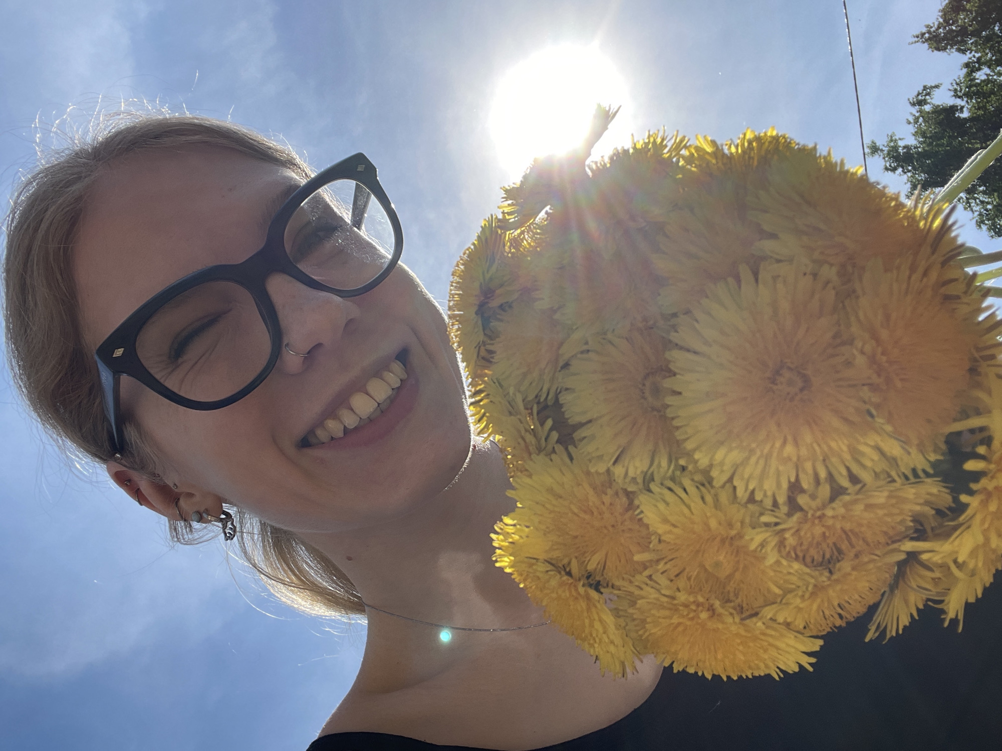A low-angle, medium shot depicts a young woman with light skin, wearing black-rimmed glasses and a nose ring. She is smiling broadly, her teeth visible. She holds a large bouquet of yellow flowers, possibly dandelions, close to the camera. The sun is directly behind the bouquet, creating a bright glare. The background includes a clear blue sky and a hint of green foliage in the upper right corner. The location cannot be determined without additional information.