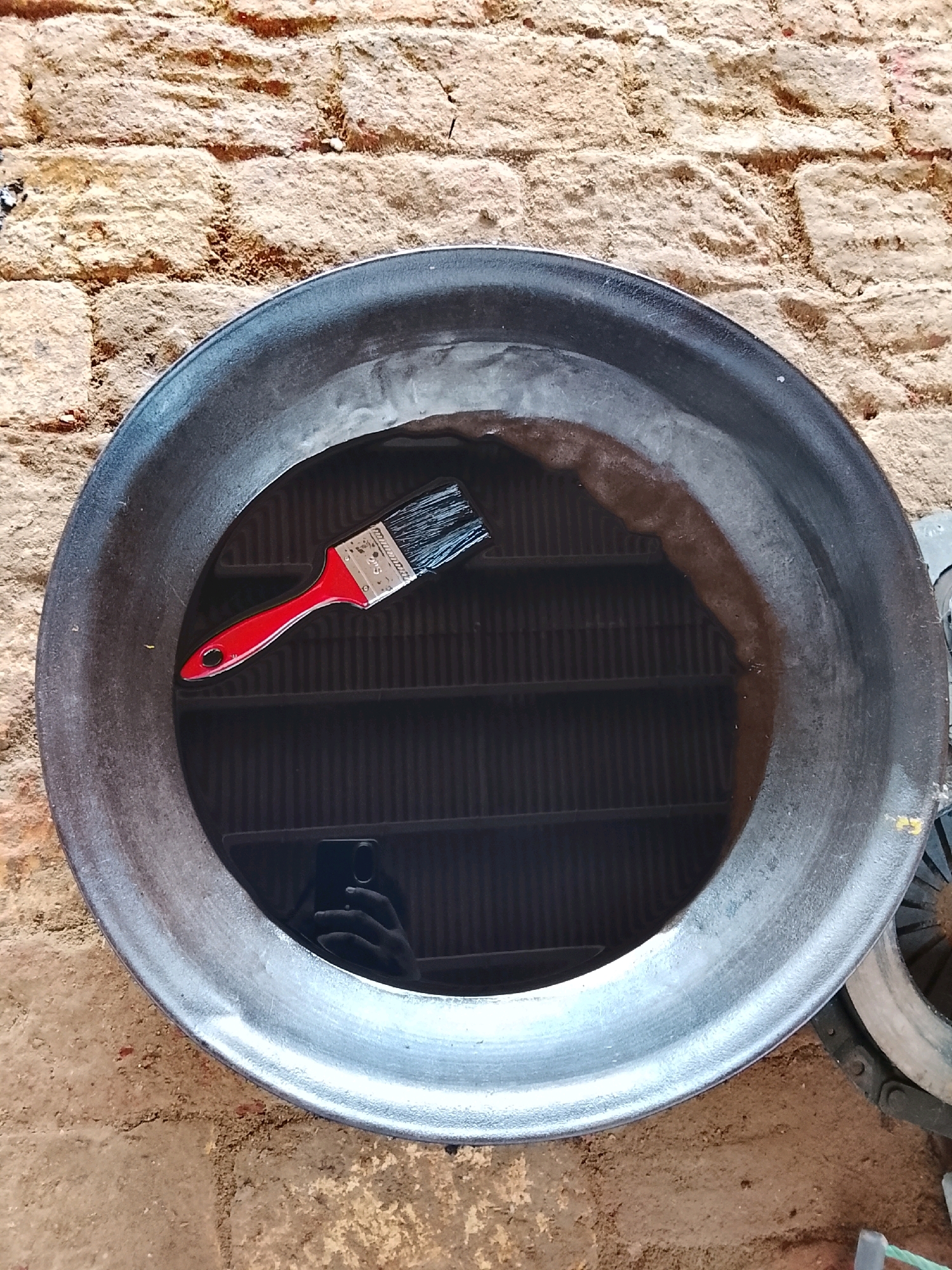 A flat, circular, dark-colored object, possibly a large pan or wok, is positioned on a rough, dirt and brick surface. Inside the object, there is a dark liquid, likely water, with a brown film on the surface near the rim. A red-handled paintbrush with black bristles is submerged in the liquid. The reflection of a person holding a smartphone is visible in the liquid. The background consists of a brick wall. The scene appears to be outdoors, with natural lighting. This location is Pataar Goth, Pakistan.