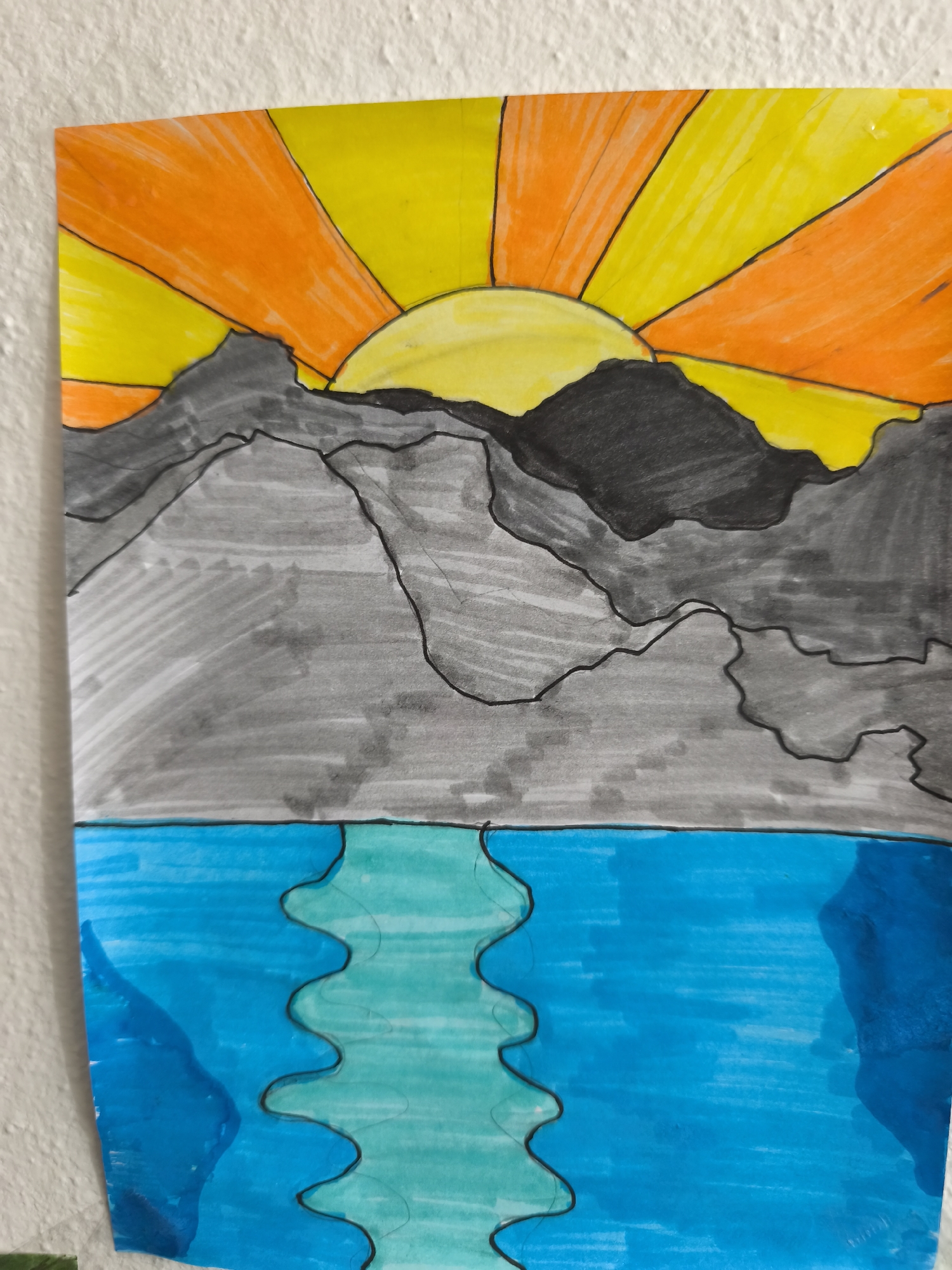 The image is a drawing of a sunrise over mountains and a body of water. The sun is depicted as a bright yellow orb with orange rays emanating outwards. Mountains, colored in various shades of gray, are layered in front of the sun. Below the mountains is a body of water, primarily colored in shades of blue and turquoise. The water has a wavy line separating two different shades of blue, suggesting reflections or ripples. The drawing is on a piece of paper attached to a textured white wall. There are no people or animals present in the drawing. The overall scene is a stylized landscape. The image does not provide any information about the city of Choceň, Czechia.