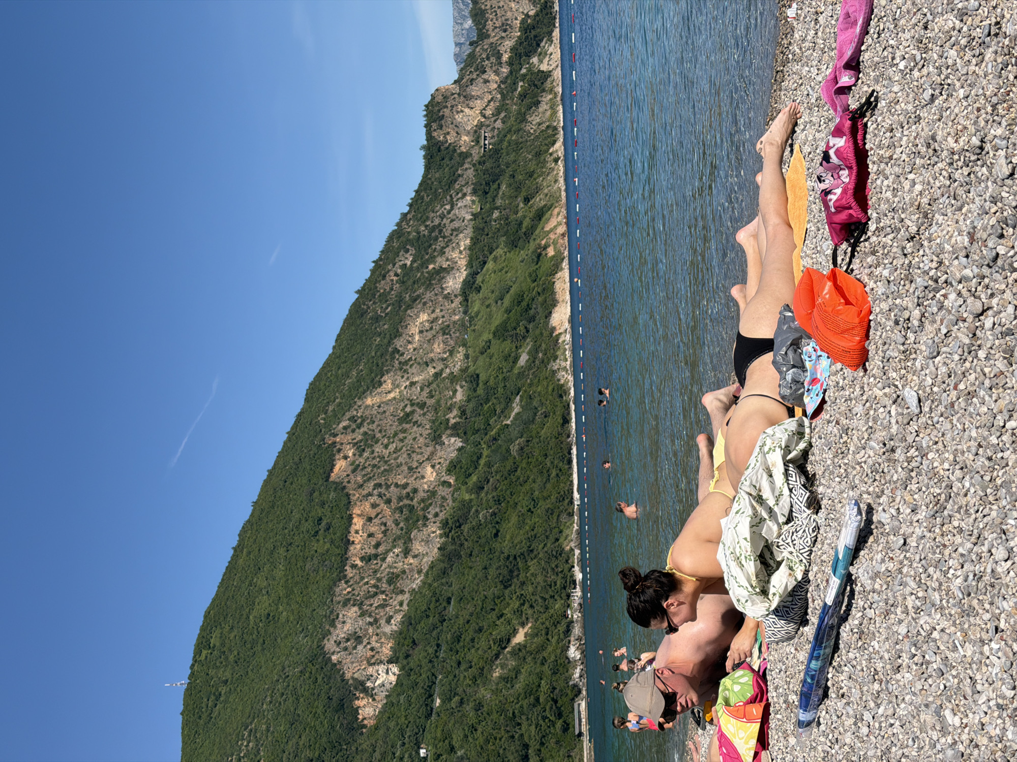 A coastal scene in Prijevor, Montenegro, shows a beach populated with people. On the right side of the image, a couple is lounging on a pebbled beach. The woman wears a yellow bikini top and black bottoms, while the man sports a tan baseball cap and sunglasses. They are surrounded by personal belongings like towels, bags, and clothing. In the water, several people are swimming, with a line of buoys indicating a swimming boundary. To the left, a steep, forested mountain rises sharply against a clear blue sky. The mountain occupies a substantial portion of the image, contrasting with the calm sea.
