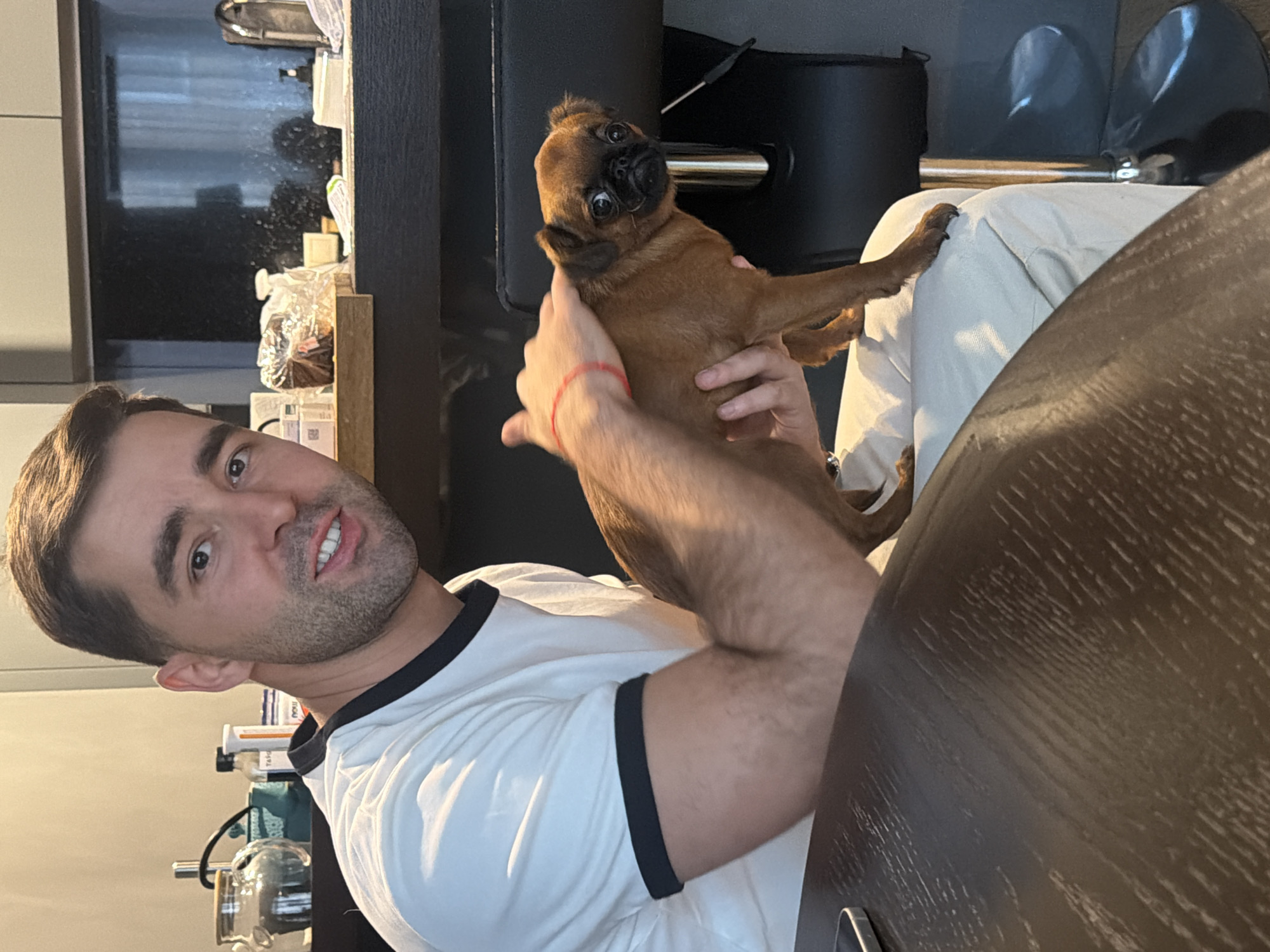 A young man, casually dressed, sits indoors holding a small, light brown dog. The man, with short dark hair and light stubble, wears a white t-shirt with black trim and a red string bracelet on his right wrist. He looks directly at the camera with a slight smile, his mouth slightly open. The small dog, possibly a Brussels Griffon breed, has short, bristly brown fur, a black nose, and large, wide dark eyes. It is cradled in the man's hands and also gazes towards the camera with a curious or slightly surprised expression.
The setting appears to be an indoor home environment, possibly a kitchen or living area. A dark wooden table is visible in the foreground on the right. In the background, various household items like bottles and jars are arranged on shelves or counters, one clearly displaying the text "NOW". A dark chair or stool with a metal component is situated behind the dog. The lighting suggests it is daytime. The image captures a candid or posed moment of affection between the man and his pet. There are no specific visual cues within the image to indicate its location as Krasnoyarsk, Russia.