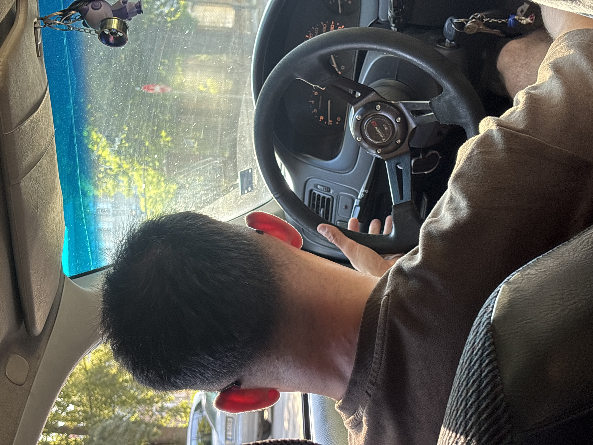 The image shows a man driving a car, seen from behind and slightly to the side. The man has short, dark hair and is wearing a brown t-shirt. He has bright red ear covers or accessories attached to his ears. He is holding the steering wheel, which has a "T-HORN" logo on it. The car's dashboard and windshield are visible. Through the windshield, trees and buildings can be seen, suggesting an urban or suburban environment. The presence of a "T-HORN" steering wheel could indicate a customized or performance-oriented vehicle. The image appears to be taken during the daytime, with sunlight filtering through the trees. No specific landmarks of Varna, Bulgaria are visible.
