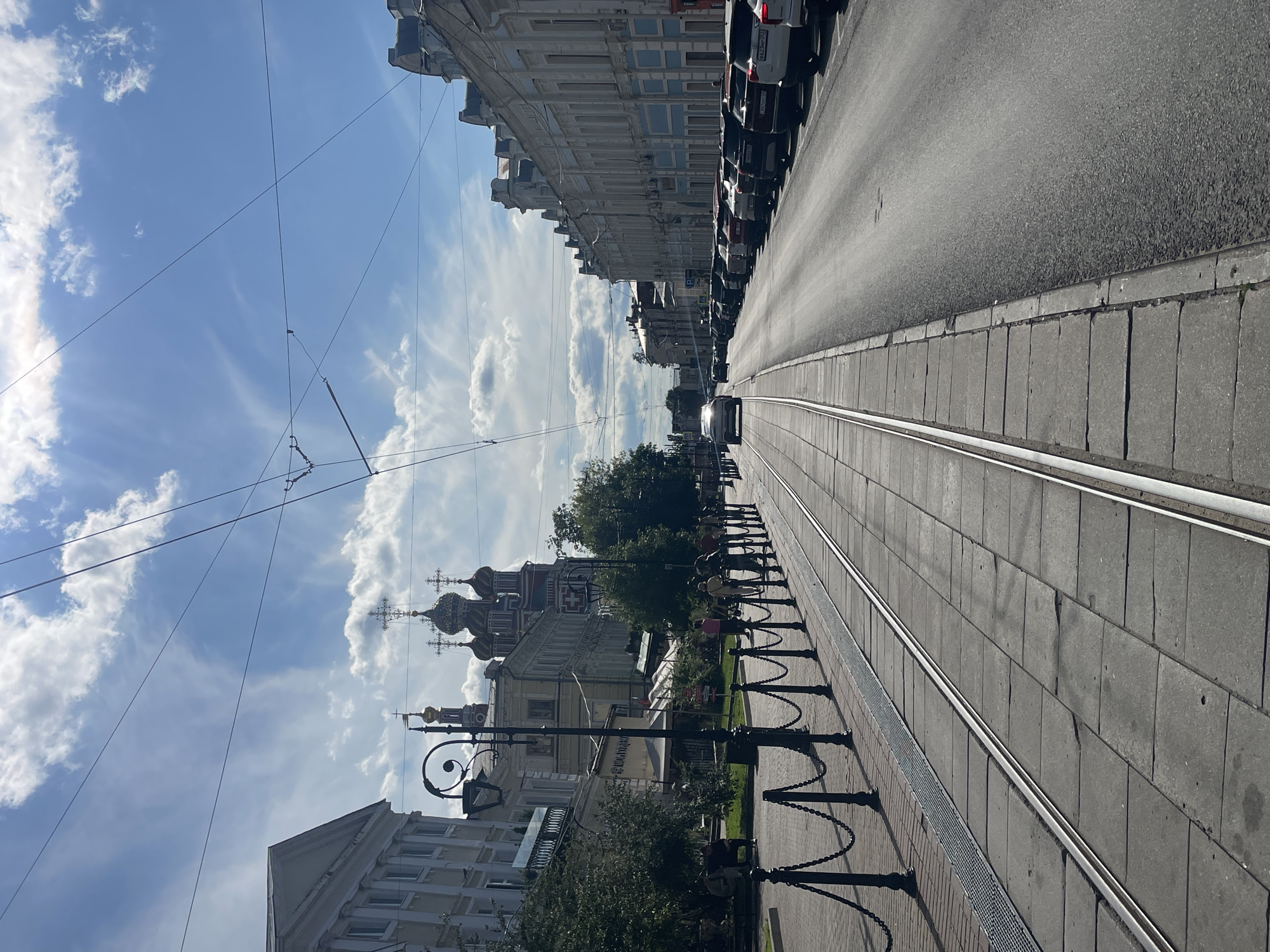 The image shows a street scene in Nizhny Novgorod, Russia, with tram tracks in the foreground. On the left side of the street, there are buildings with classical architecture, trees, and people walking along the sidewalk. A chain fence separates the sidewalk from the tram tracks. On the right side of the street, there are more buildings and a row of parked cars. The sky is blue with scattered clouds, and there are overhead tram wires. The overall impression is of a bright, sunny day in a historic urban environment.