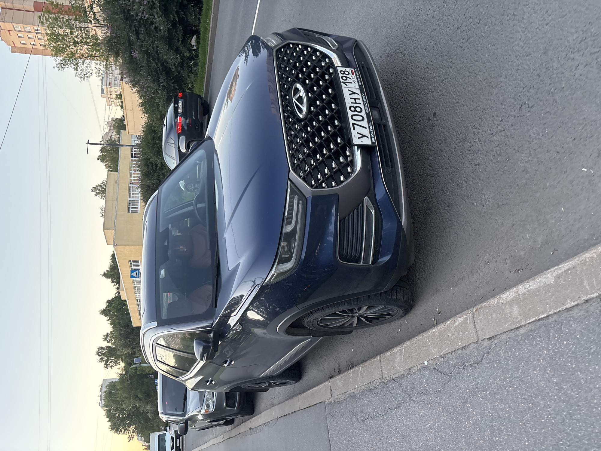 A dark blue Chery Tiggo 8 Pro is parked on the right side of a two-lane road, close to the curb. The car is facing slightly away from the camera, with its front end visible. The license plate reads "Y708HY 198". Behind it, a gray SUV is also parked. In the background, there are buildings, trees, and power lines. The sky is clear and pale. The scene appears to be an urban environment.