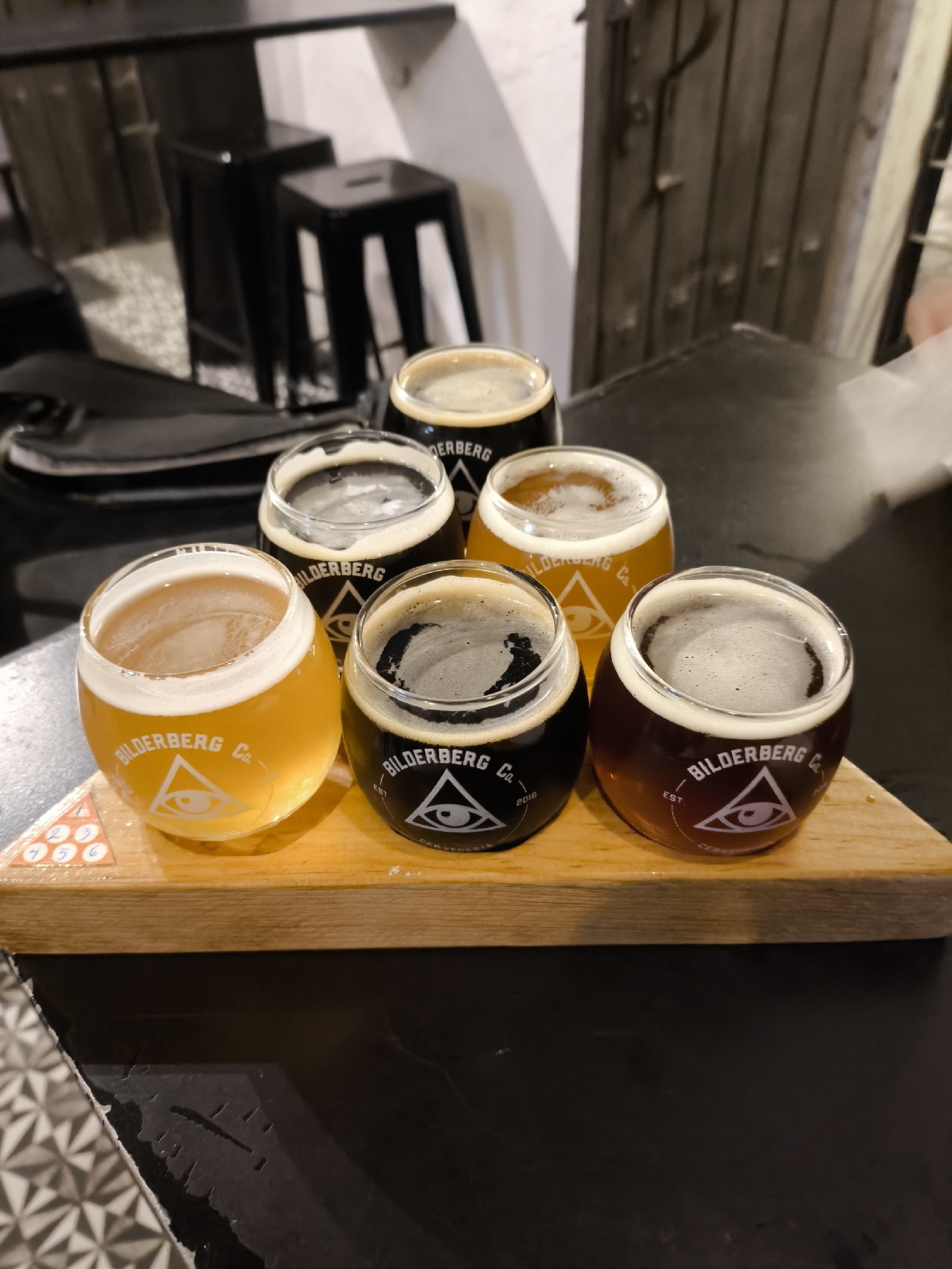 The image shows a wooden serving tray containing six small glasses, each filled with different types of beer. The glasses have a consistent branding: "BILDERBERG Co." along with a logo of an eye inside a triangle, suggesting they come from the same brewery or establishment. The beer colors range from light yellow to dark brown and black. The tray also features small, numbered triangles, possibly for differentiating the beers.
The setting appears to be a bar or restaurant with black tables and chairs. The patterned floor is visible. The location is in the city of Puebla, Mexico.