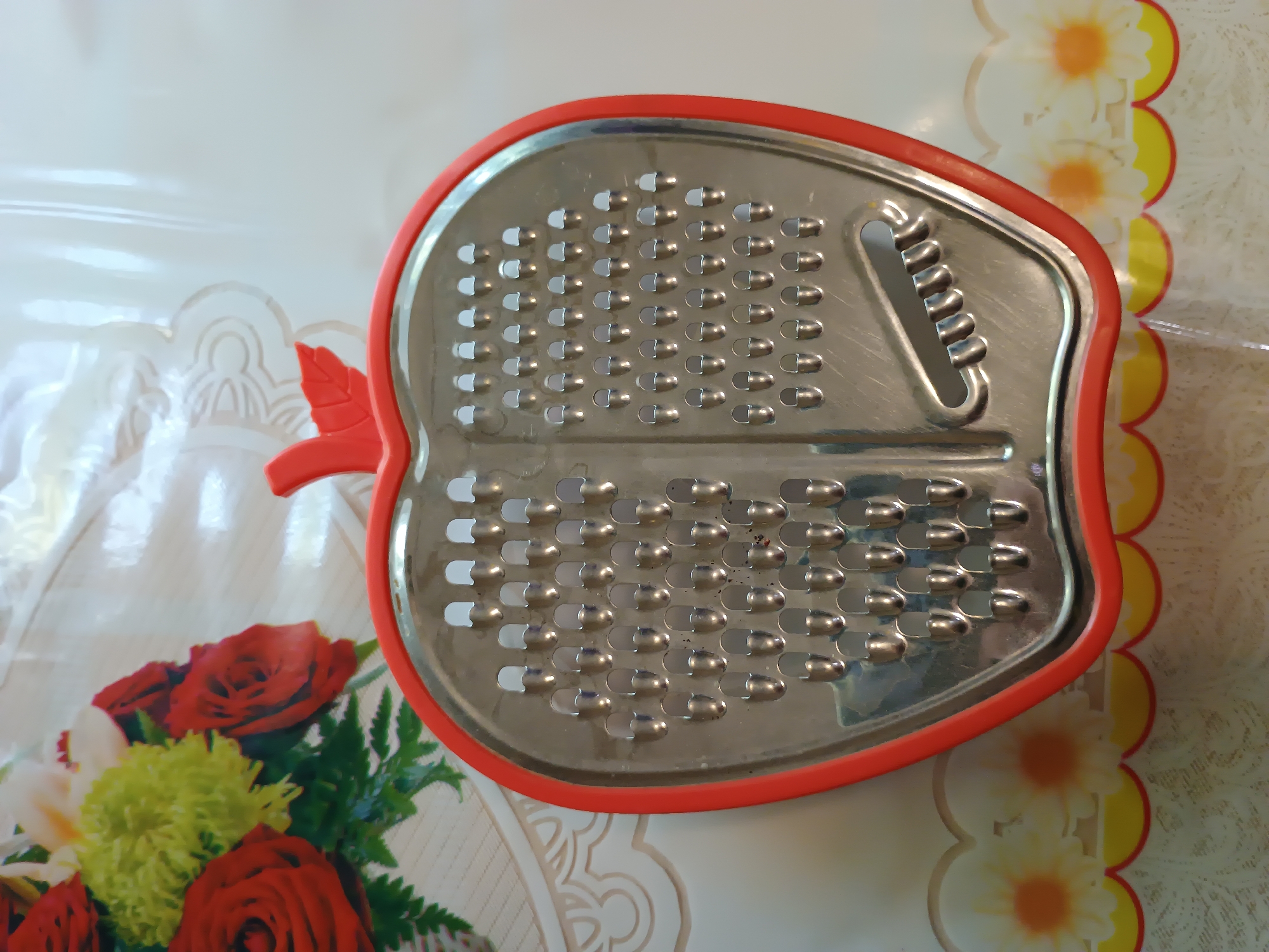 A close-up, eye-level shot shows a red, apple-shaped grater with a metal grating surface, placed on a tablecloth. The tablecloth has a floral pattern with white and yellow flowers against a light background, with a red and yellow scalloped border along the bottom. In the upper left corner of the image, a bouquet of red roses and green foliage is partially visible. The grater itself is the primary subject, with its distinct shape and the rows of sharp grating holes clearly visible. There are no people or visible actions taking place in the image. The scene is indoors, with soft lighting.