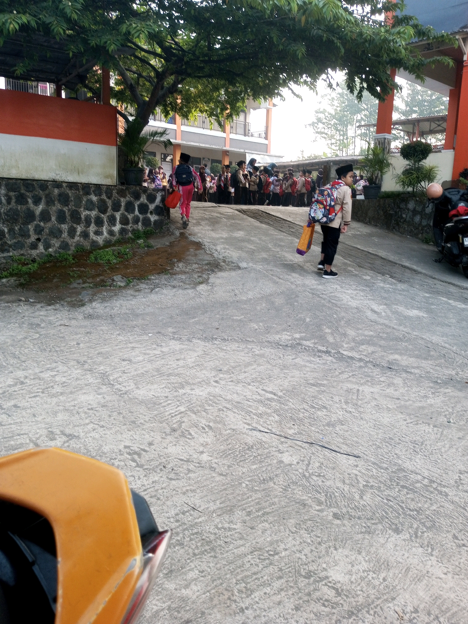 In Krasak, Indonesia, a school environment is depicted. A group of students, dressed in uniforms, are gathered outdoors on a paved pathway in front of a building with orange and white accents and a stone wall. Two students are walking away from the camera, one in pink pants and a backpack, carrying an orange bag, and another in a tan jacket, black pants, and a colorful backpack, also carrying an orange bag. A motorcycle is parked to the right of the frame. Trees with lush green foliage overhang the area. The scene suggests a typical school day with students arriving or departing.