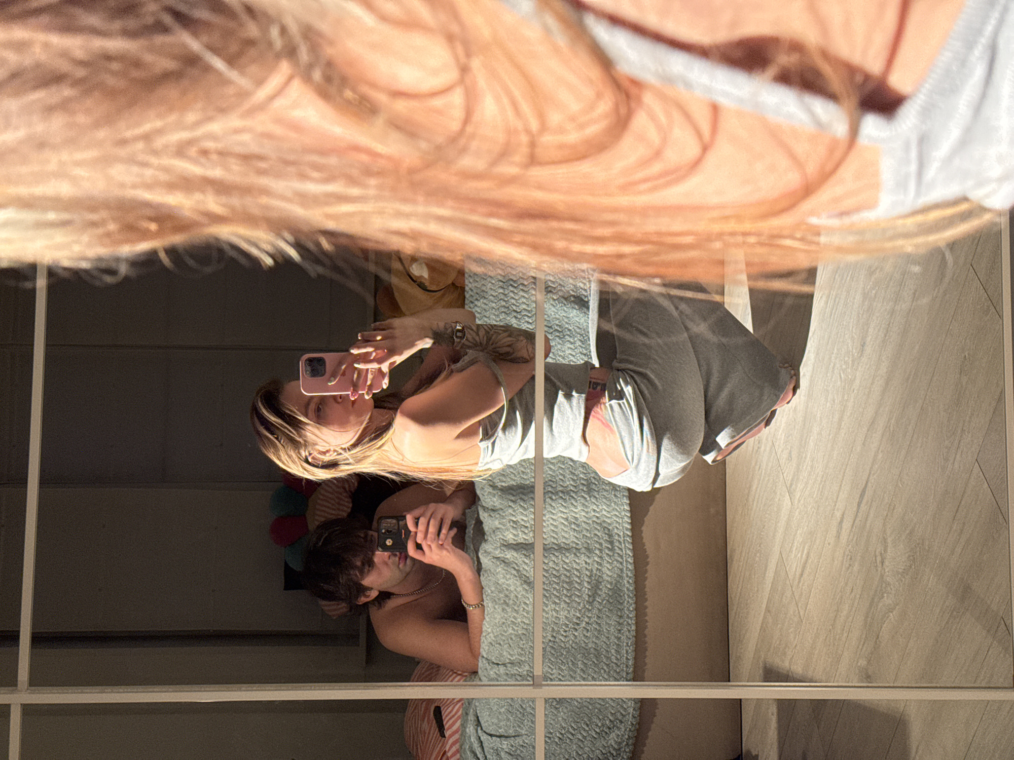A young couple is taking selfies in what appears to be a bedroom. A man, shirtless with a necklace, is lying on a bed and holding a smartphone, looking at the screen. Beside him, a woman is crouching and holding a smartphone up to take a selfie in a large mirror that covers the wall. She is wearing a light gray tracksuit. The room is lit by sunlight coming from the left, casting shadows on the wall and floor. The floor is made of light-colored wooden planks. The bed has a textured, light blue comforter. In the background, on the bed, there are some colorful stuffed pillows. The overall scene is captured from a low angle.