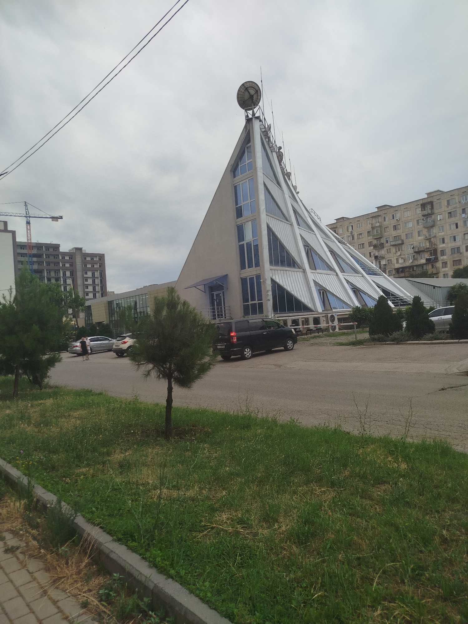 The image depicts a street scene in Rustavi, Georgia, characterized by a dramatic, angular building with a large satellite dish on its roof. The building's facade features a series of large, triangular windows set against a light-colored, possibly metallic, frame. To the right of this structure is a multi-story apartment building with numerous windows, typical of Soviet-era architecture. In the foreground, a paved sidewalk runs alongside a strip of grass and a road. Several cars are visible on the road, including a black SUV and a silver sedan. A construction crane and an unfinished building are present in the background, suggesting ongoing development in the area. The sky is overcast with a neutral gray hue. The scene appears to be a daytime, possibly late afternoon, snapshot of an urban environment.