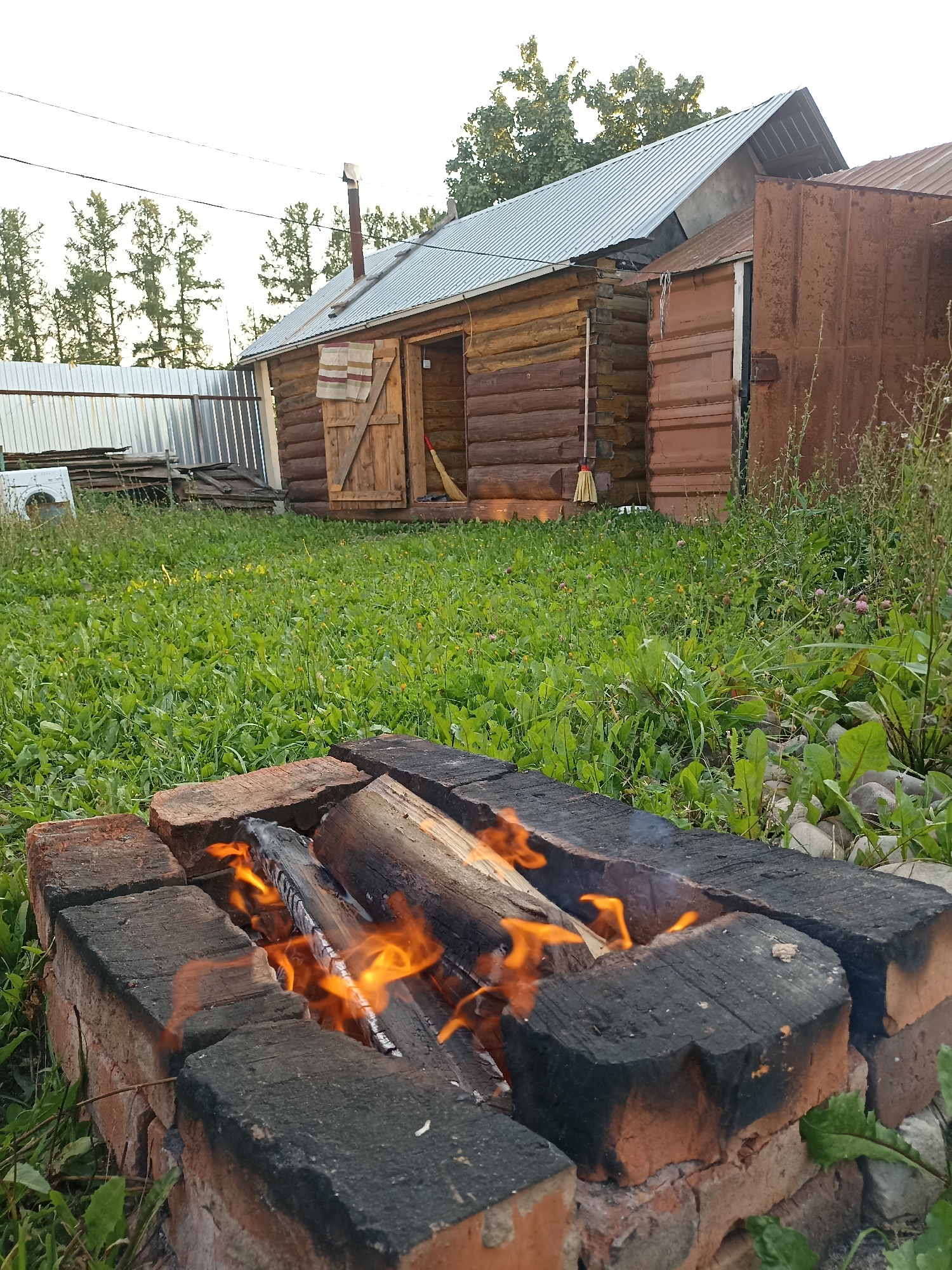 In the village of Maldykasy, Russia, a small outdoor fire burns in a brick fire pit, with logs stacked inside. The fire pit is situated in a grassy yard, with a wooden log cabin visible in the background. The cabin has a metal roof and a wooden door. A broom leans against the side of the cabin. To the left of the cabin, a corrugated metal fence encloses a section of the yard, and a washing machine is visible behind it. The sky above is overcast.