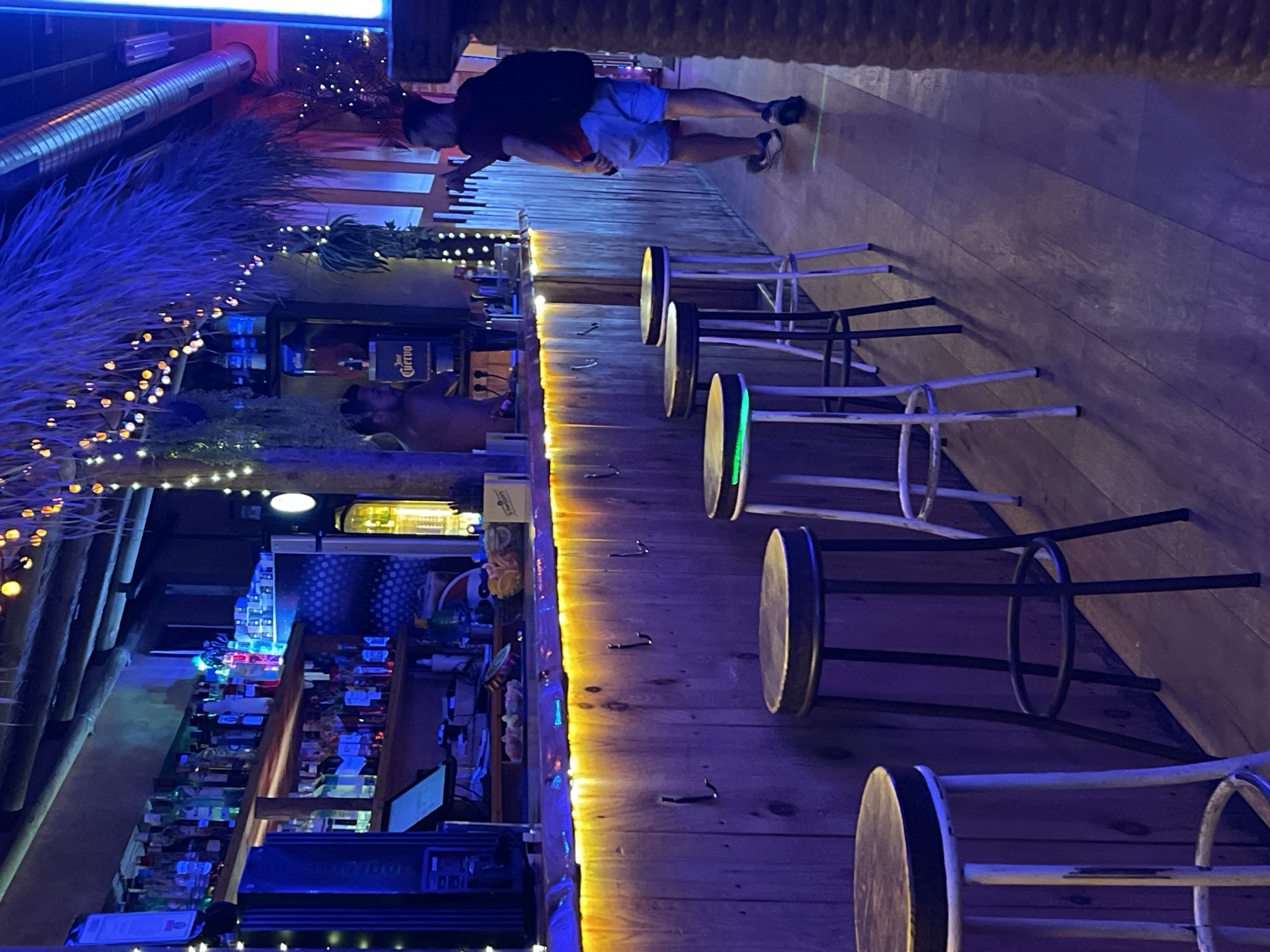 The image depicts the interior of a bar in Barcelona, Spain, illuminated by blue and yellow neon lights and strings of small, warm-toned lights. The bar has a rustic wooden facade with several hooks. A wooden counter is lined with stools, some of which have illuminated green rings around their seats. Behind the counter, shelves are stocked with numerous bottles of alcohol, and a large dispenser is visible. Two men are present; one appears to be a bartender, standing behind the counter, while the other, wearing a red shirt and light blue shorts, is walking away from the camera, looking towards the right side of the frame. The floor is made of polished wood. The atmosphere suggests a lively evening establishment.