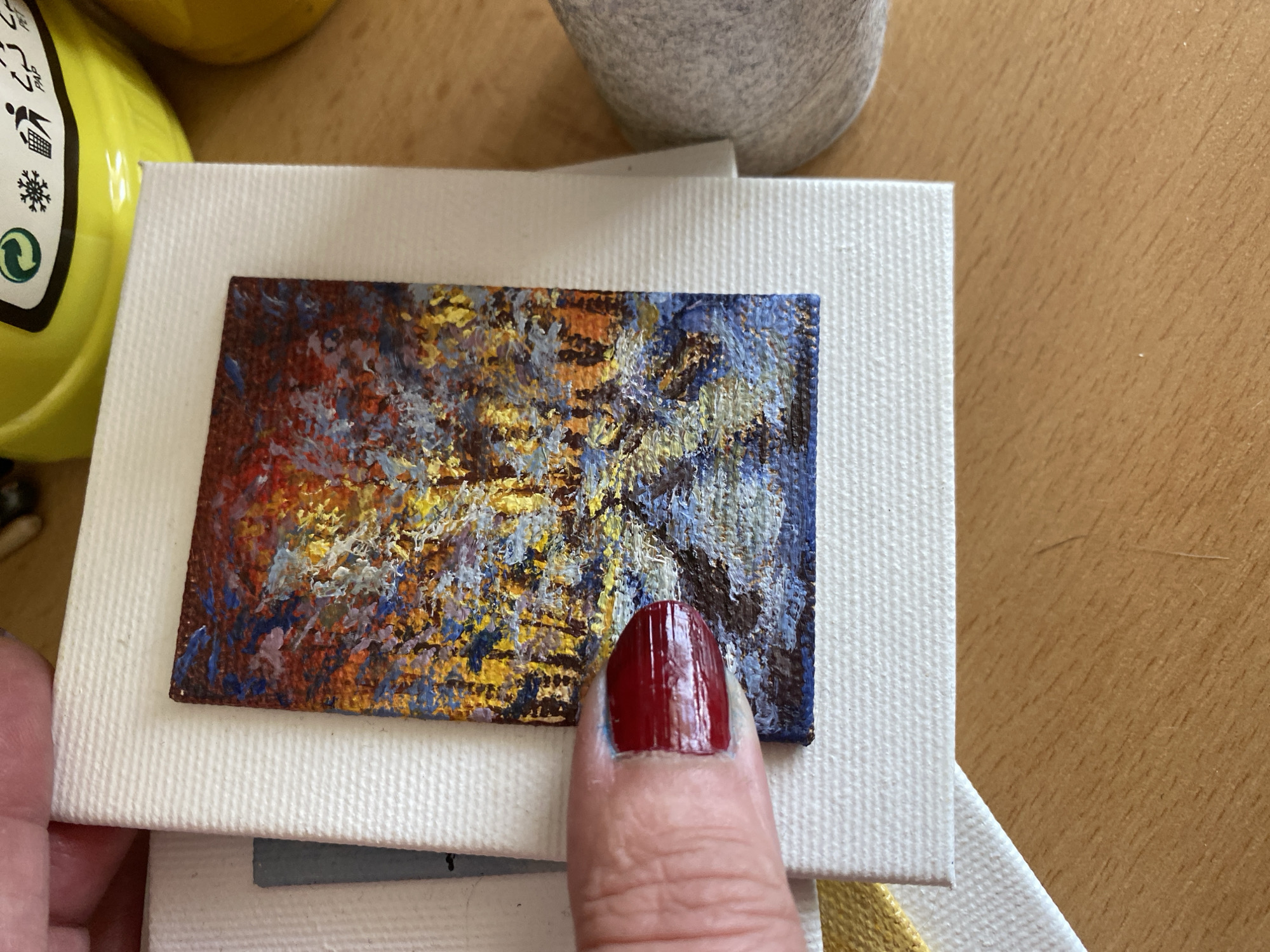 A close-up, high-angle shot focuses on a miniature painting, approximately 2x3 inches, depicting a colorful, abstract landscape with trees and water. The painting is placed on a larger white canvas, which in turn rests on a light brown wooden surface. The painting's texture is visibly thick with brushstrokes. A person's thumb, with dark red nail polish, gently touches the edge of the miniature artwork. In the background, out of focus, a yellow container with recycling symbols and a grey, pebble-like object are visible. The lighting suggests an indoor setting. The image is not specific to Bílina, Czechia, or any other location.