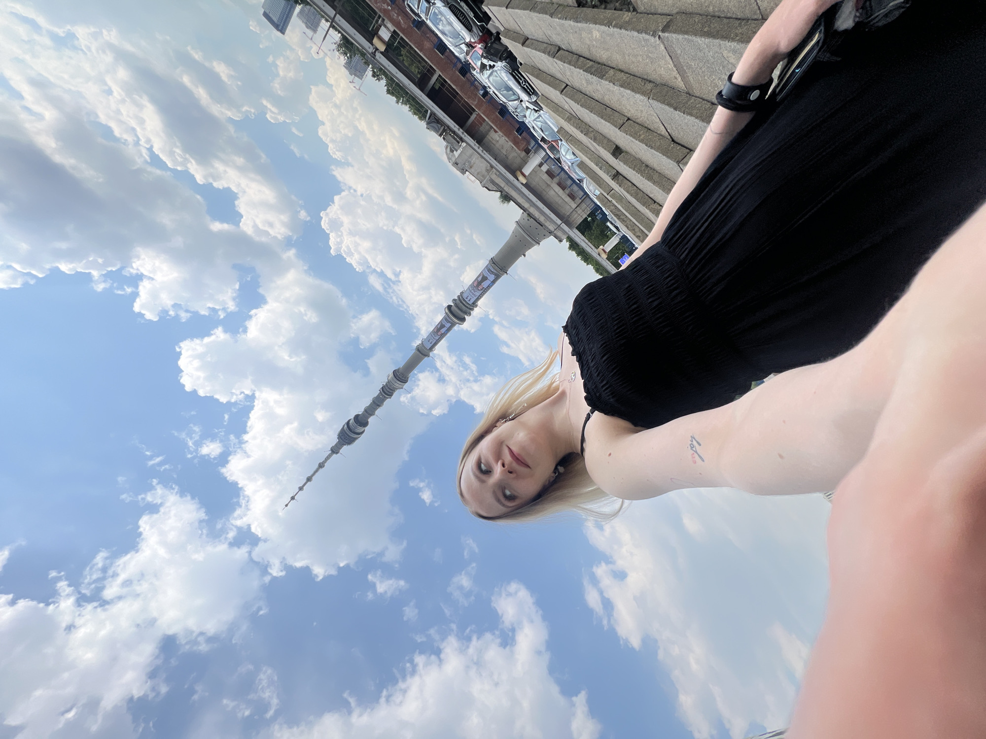 A young woman with blonde hair, wearing a black dress, is taking a selfie outdoors with a tall tower and a bright blue sky with white clouds in the background. She is looking up and to her left, with a slight smile. The tower, which appears to be the Ostankino Tower, is visible in the upper half of the image, extending from the middle towards the top right corner. The lower part of the image shows a textured stone staircase and a line of parked cars. The overall setting suggests an urban environment with a prominent landmark.
