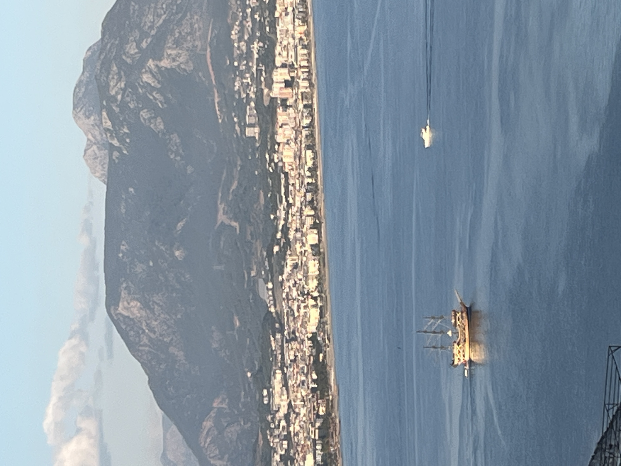 The image shows a coastal scene in Alanya, Turkey. A large, dark mountain rises along the coast, adjacent to a densely built-up urban area with closely packed buildings. The sea occupies a significant portion of the frame. A historical sailing ship with masts and rigging is sailing on the water, appearing illuminated by the sun. Further in the distance, another smaller watercraft is visible. Part of a structure with bars is visible in the lower right corner, suggesting an elevated viewing point.