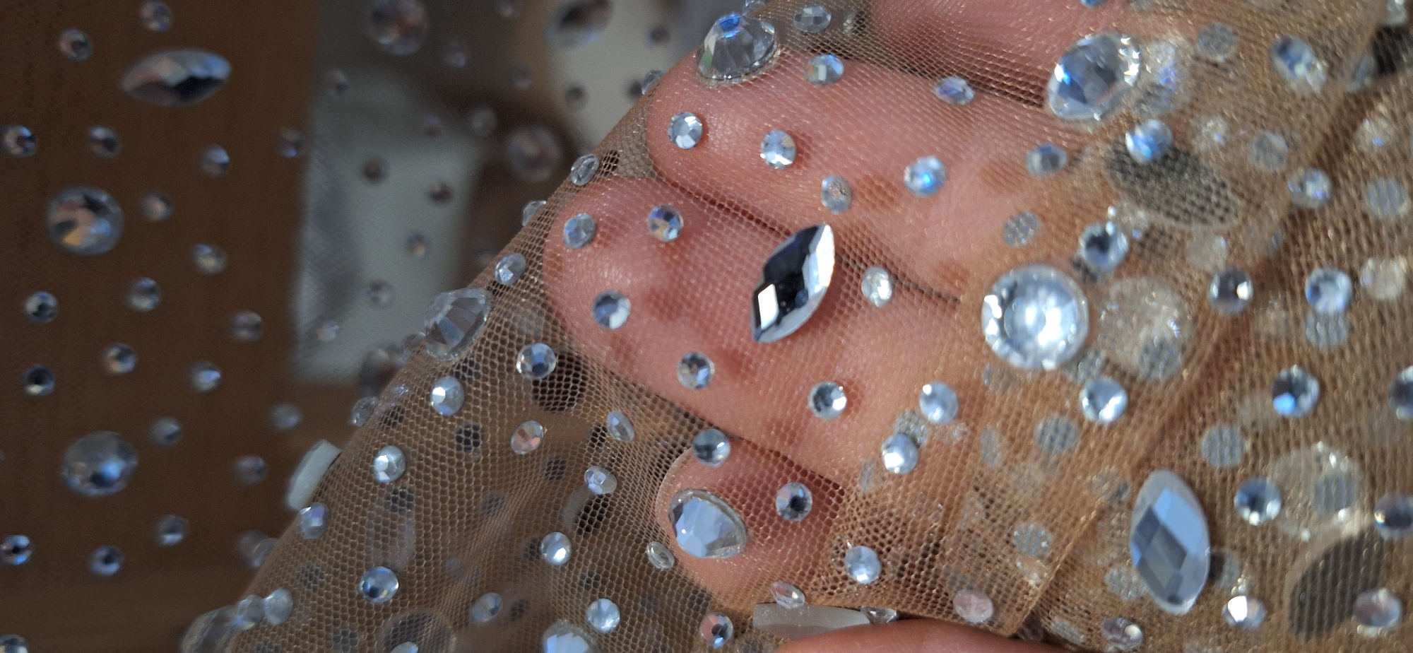A close-up shot reveals a hand holding up a section of sheer, beige-toned fabric. The fabric is adorned with numerous clear rhinestones of varying sizes and shapes, including round and teardrop cuts. The hand is visible through the mesh of the fabric, with the rhinestones appearing scattered across its surface. The background is blurred and indistinct, obscuring any specific details about the location. There is no indication that the location is in Blagoevgrad, Bulgaria.