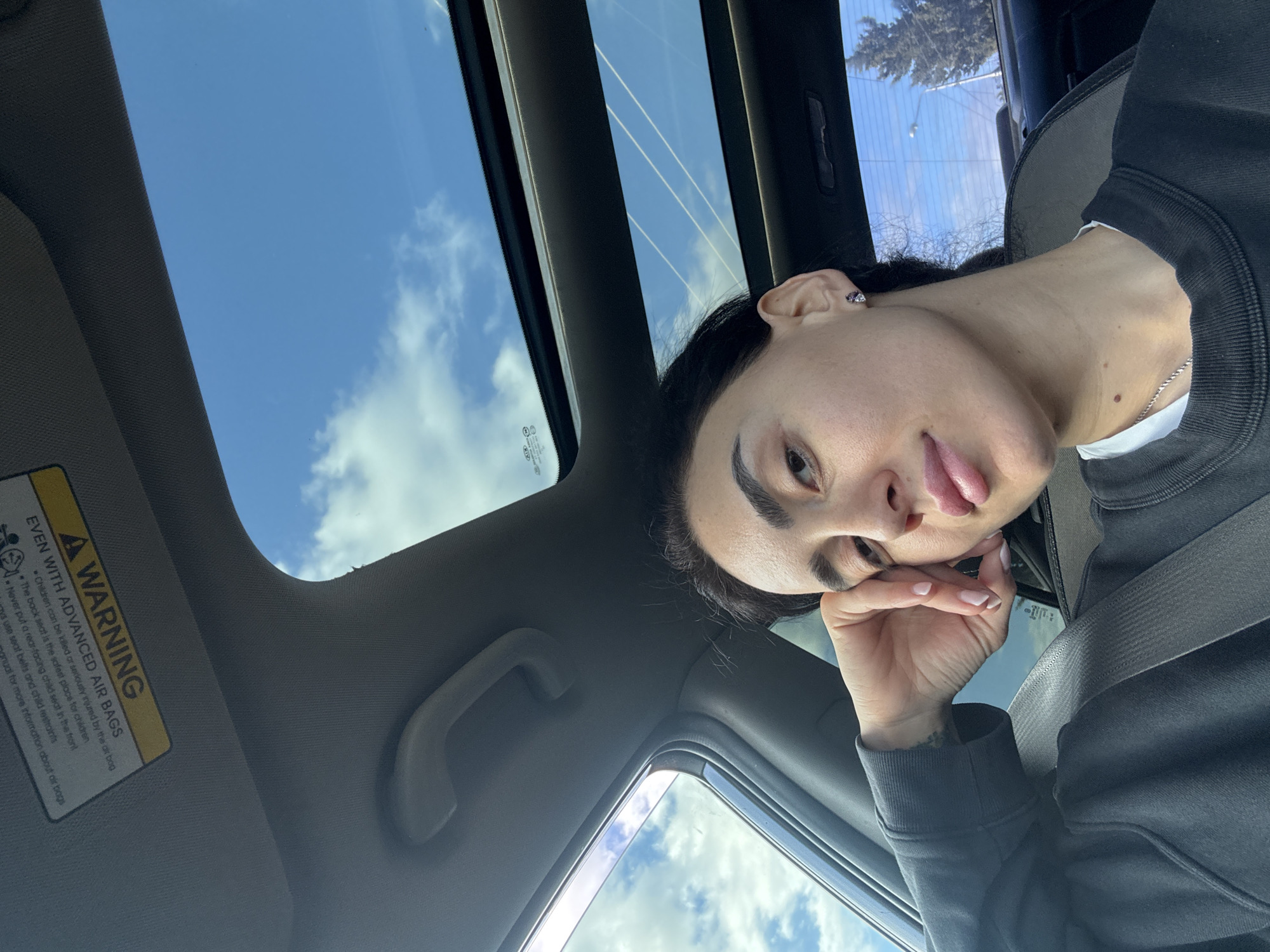 The image shows a woman inside a car, likely in a city environment. She's wearing a dark sweatshirt and has her seatbelt on. The car has a sunroof, providing a view of the sky. The woman is resting her head on her hand and sticking her tongue out slightly. The sun visor on the driver's side is down, displaying a warning label about airbags. Through the window and sunroof, glimpses of buildings and power lines suggest an urban area. There is no evidence to determine if the location is in Tbilisi, Georgia.