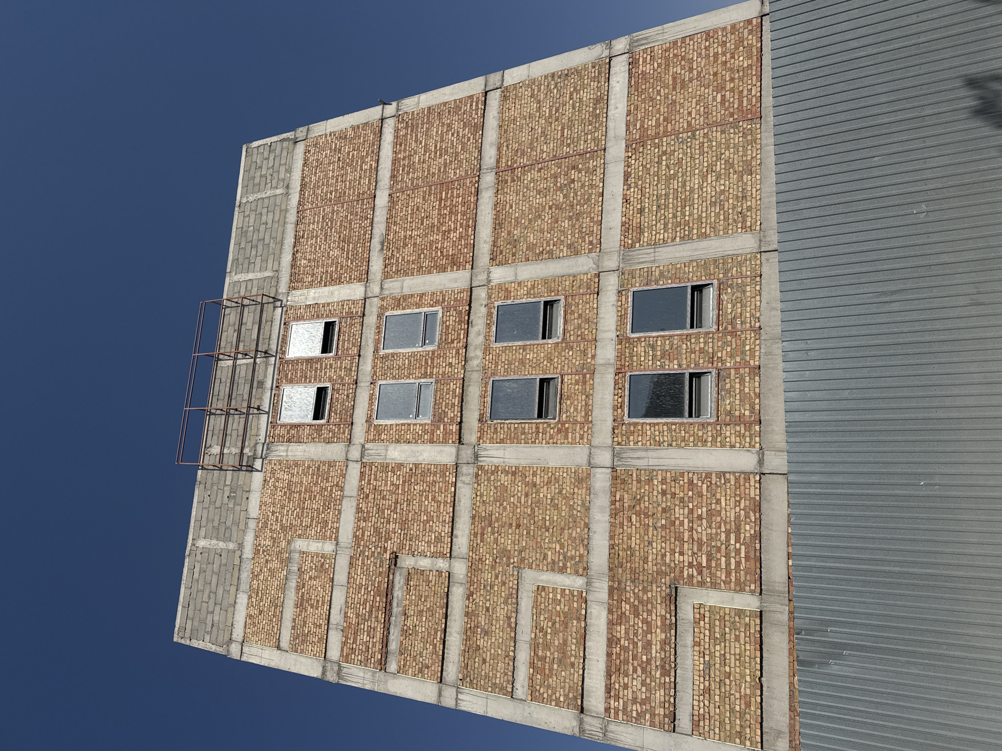 The image shows a close-up, low-angle view of the exterior of a multi-story building under construction, against a clear blue sky. The building's facade is a combination of brickwork and concrete. The brickwork is composed of small, yellowish-brown bricks, laid in a stretcher bond pattern. Concrete elements, such as horizontal bands and vertical columns, frame sections of the brickwork.
Several windows are visible on the facade, appearing as dark rectangles with reflective glass, suggesting they are either tinted or covered. Some of these windows have visible frames and are open at the top. The top of the building features a section of grey concrete blocks, with exposed rebar and a rudimentary metal railing, indicating ongoing construction or unfinished work.
The lower portion of the image is dominated by a corrugated metal fence, which runs horizontally across the frame, obscuring the base of the building. The fence is a dull metallic gray and has a ribbed texture. Shadows of foliage are visible on the fence in the bottom right corner, suggesting the presence of trees nearby. The overall impression is of an unfinished urban structure.
