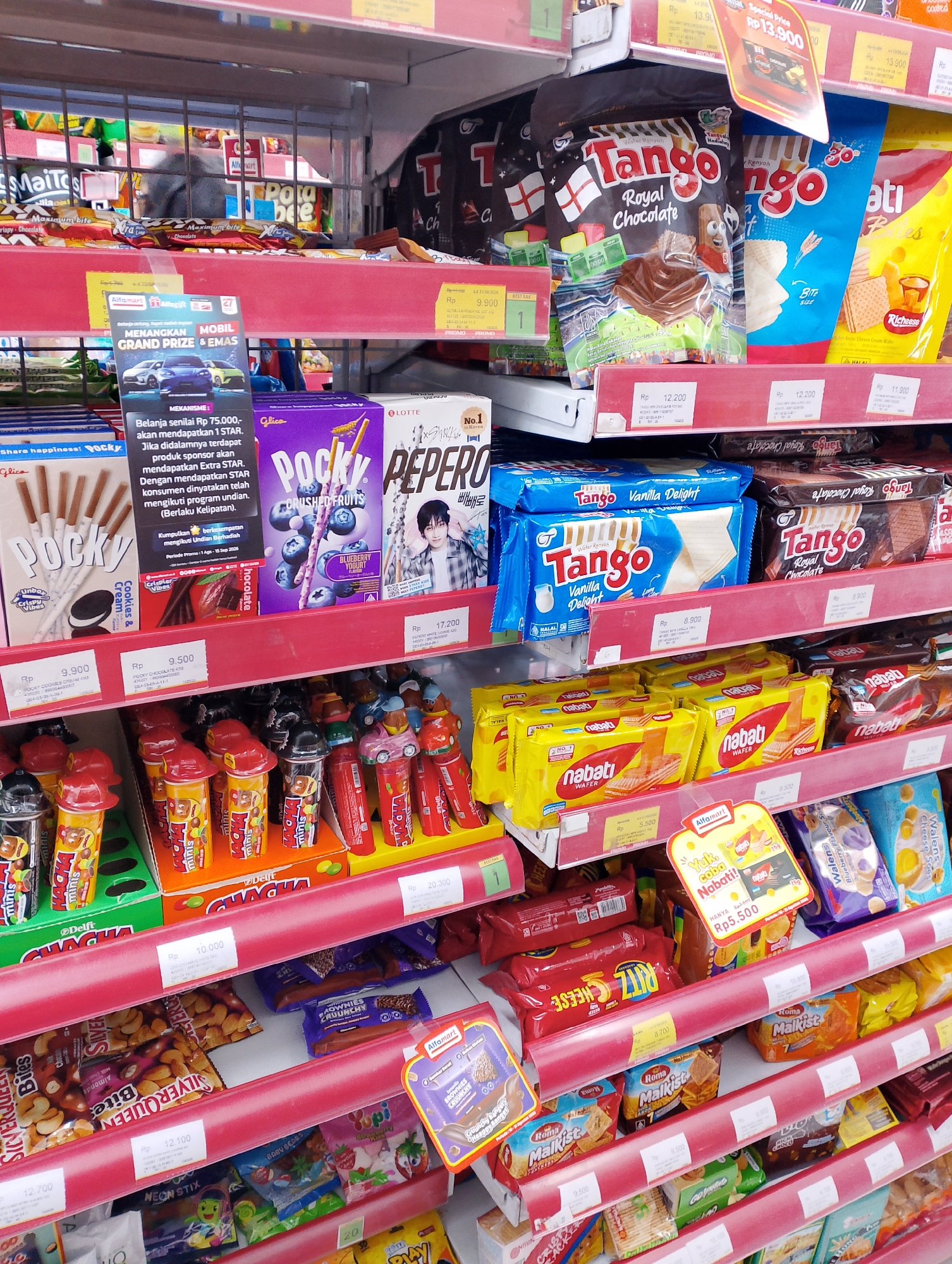 This is a medium-angle, eye-level shot of a brightly lit, well-stocked shelf in a convenience store, likely an Indomaret or Alfamart, in Magelang, Indonesia. The shelves are filled with various packaged snacks and sweets, predominantly cookies, wafers, and candies. The products are arranged in neat rows, with prices displayed on red shelf tags.
Prominently featured are several brands of wafers, including Tango in "Royal Chocolate" and "Vanilla Delight" flavors, and Nabati wafers in yellow packaging. Other visible items include Pocky sticks in "Cookies & Cream" and "Blueberry Yogurt" flavors, and a package of Pepero cookies. There are also small, individually wrapped candies and snacks, some in colorful packaging with cartoon characters.
A promotional sign for a "Grand Prize" with a car and gold imagery is displayed on the top shelf, suggesting a promotional event. The overall impression is one of abundance and consumer choice within a typical retail environment. The store appears clean and organized, with no visible customers in the immediate vicinity, though a blurry figure can be seen on a higher shelf in the background. The lighting is bright, casting minimal shadows and illuminating the products effectively.