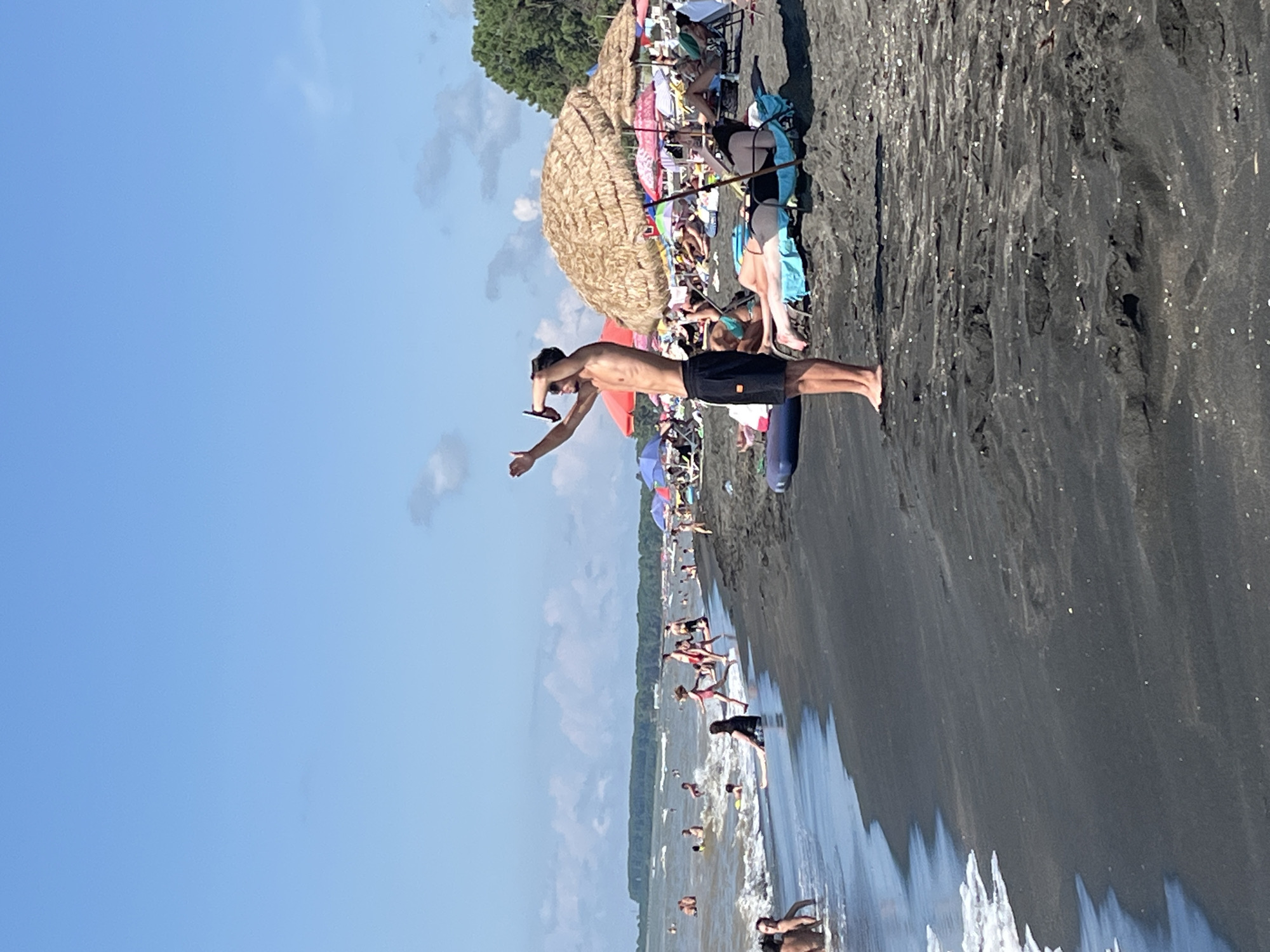 This image captures a crowded scene at Ureki, Georgia's famous magnetic sand beach on a sunny day. The foreground features dark, wet sand with small white particles, indicating the presence of magnetic sand. The water's edge shows foamy waves breaking.
In the mid-ground, numerous people are engaged in beach activities. Some are swimming or wading in the moderately wavy, brown-tinted water. Others are sunbathing on loungers, many of which are shaded by straw umbrellas and colorful beach umbrellas. Several individuals are standing and walking along the shoreline.
A prominent figure in the mid-ground is a young man standing on the wet sand, wearing black shorts. He is holding a phone to his ear with his right hand, as if making a call or taking a picture, and his left arm is raised. Behind him, more beachgoers are visible, as well as a dense line of trees and foliage forming a backdrop along the coast. The sky is a clear, bright blue with a few scattered white clouds. The overall atmosphere is one of a busy, recreational beach environment.