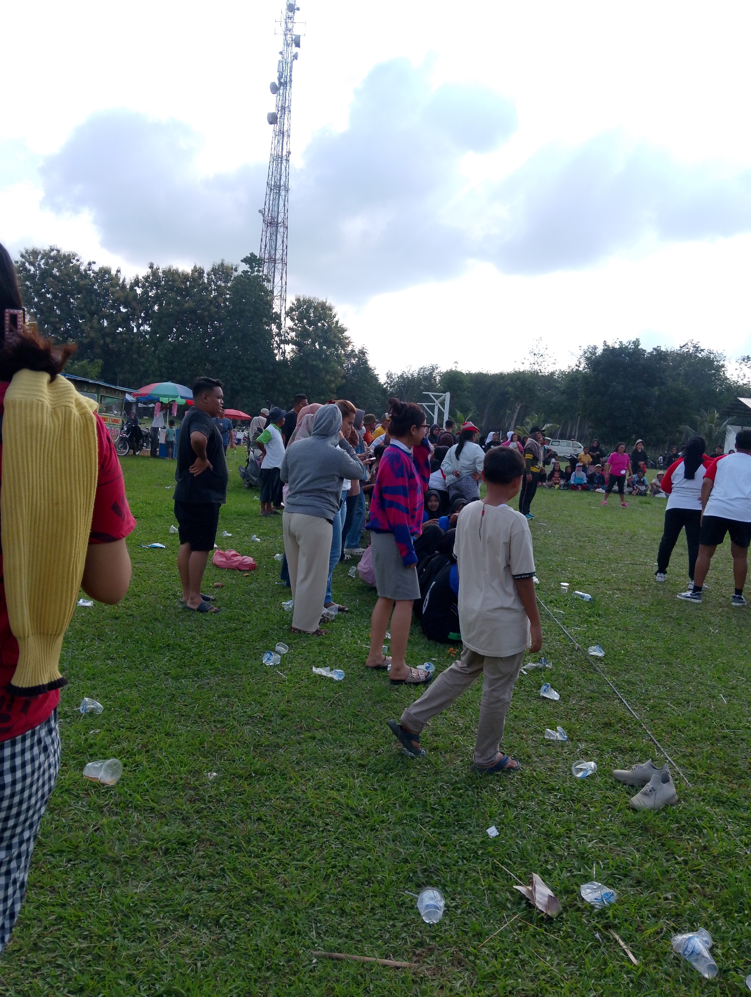 In the city of Serbelawan, Indonesia, an outdoor gathering is taking place on a grassy field, with a tall telecommunications tower visible in the background against a cloudy sky. Numerous people of various ages are present, some standing and observing, while others appear to be engaged in activities or conversing. Several food stalls or vendors are set up in the middle ground, and motorcycles are parked nearby. The ground is littered with plastic bottles and other debris, suggesting a recent event or ongoing casual use of the space. The overall impression is of a community event or informal gathering.