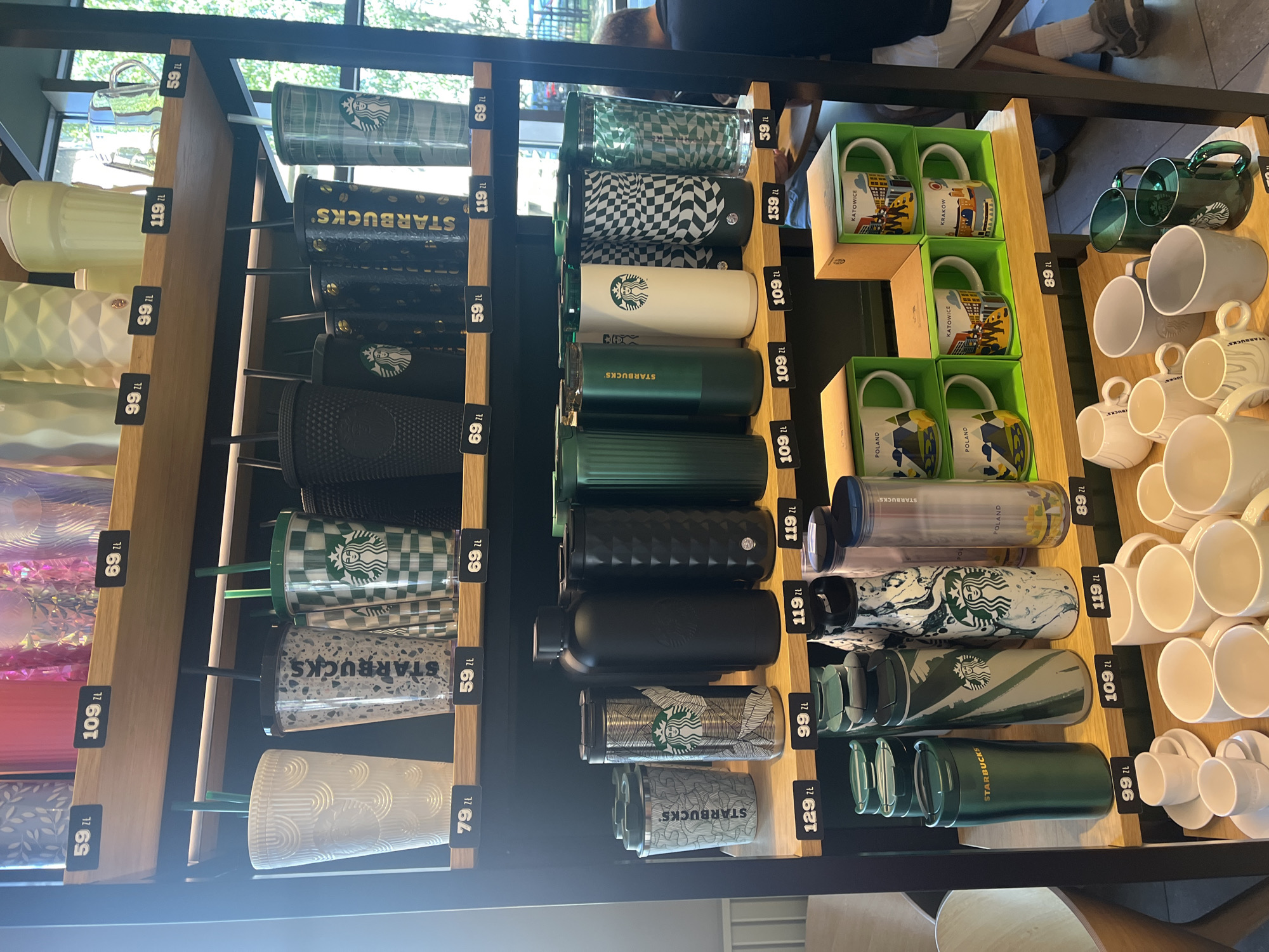 The image shows a retail display of Starbucks merchandise, primarily reusable cups and mugs, arranged on wooden shelves within a store. The merchandise is diverse, featuring various colors, patterns, and sizes, including tumblers, travel mugs, and larger cups with straws. Prices are clearly labeled below many of the items. The background suggests an indoor retail environment with subtle lighting. There is a partial view of a person sitting in the background, facing away from the camera. The setting appears to be a Starbucks store or a similar retail space selling branded merchandise.