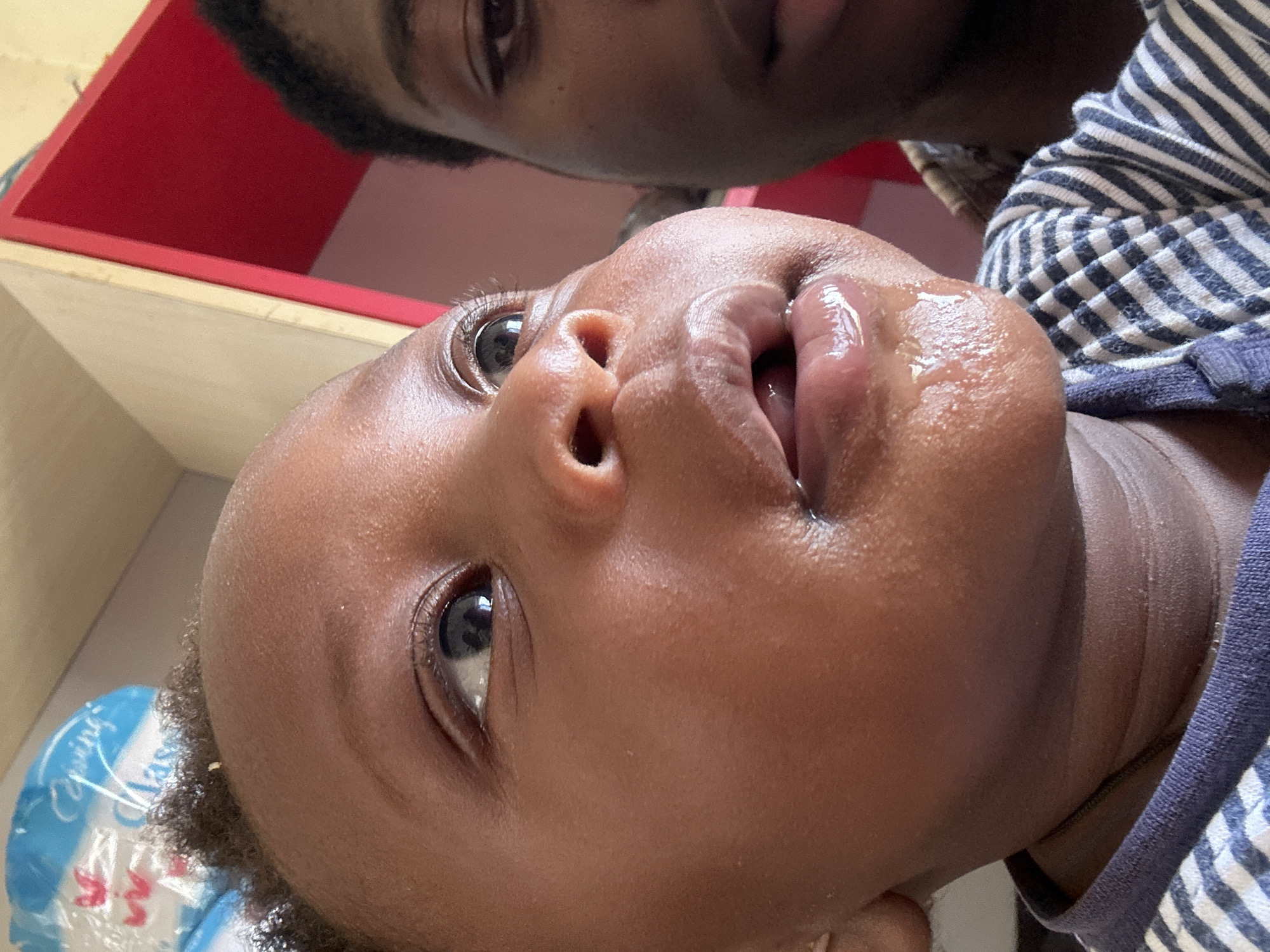 The image presents a close-up, low-angle perspective of a baby or toddler looking upwards, interacting with an adult whose face is partially visible in the background. The scene likely takes place indoors, given the visible furniture elements.
The primary subject is a young child with dark skin, dark curly hair, and wide, curious dark eyes gazing upwards. Their mouth is slightly open, revealing a wet, glossy appearance on their lips, suggesting the presence of drool or liquid, possibly from feeding or playing. The child is wearing a blue and white striped long-sleeved top.
Above and behind the child, another person with dark skin and dark hair is visible, primarily their eyes and forehead, looking down towards the child. This suggests an intimate interaction or a moment of care.
The background shows indistinct indoor elements: a red shelf or surface in the upper left and a lighter wooden surface beneath it. In the bottom left corner, part of a blue and white plastic bag or container is visible, with the word "Spring" and part of the word "Class" discernable on it.
The lighting is bright, indicating daytime or a well-lit interior. The image captures a tender, focused moment between the two individuals, possibly in Gura Top, Nigeria, as suggested by the context.