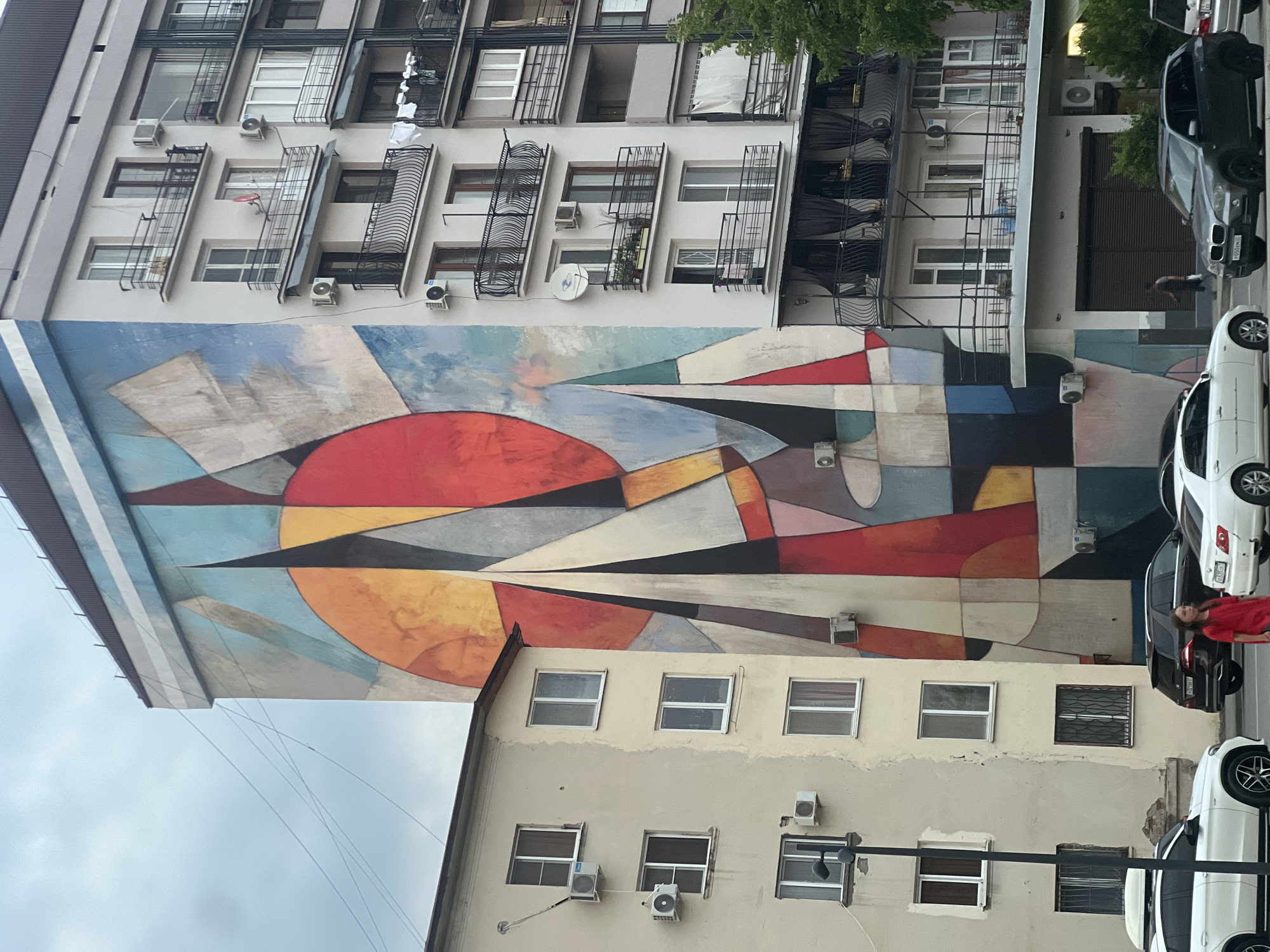 The image shows a large, colorful mural on the side of a tall, modern apartment building in Sokhumi, Abkhazia. The mural features abstract geometric shapes in vibrant colors like red, orange, yellow, blue, and gray, resembling sails or a sunburst. Adjacent to this building is a smaller, older, beige apartment building with several windows and air conditioning units. A street runs in front of the buildings, with several cars parked and driving on it, including a white SUV, a black SUV, and a white sedan. A young woman in a red dress is walking on the sidewalk. The sky is overcast.