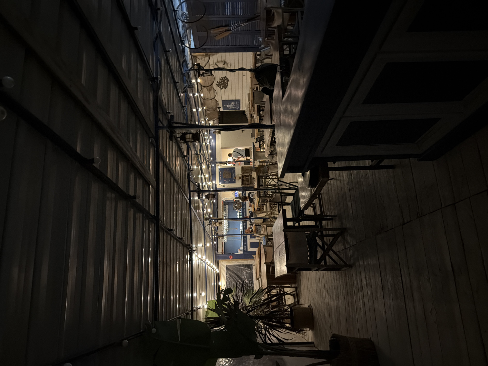 The image depicts the exterior of a restaurant in Torreilles, France, at night. The scene is illuminated by string lights and lanterns hanging from the corrugated metal roof. The restaurant features wooden tables and chairs arranged on a wooden deck. In the foreground, a bar area is visible with a dark countertop and paneled base. Towards the back, there are people seated at tables, and a man in a white shirt and black pants is standing near a doorway. The restaurant appears to have a nautical or beach theme, with some decorative elements suggesting this, such as oars mounted on the wall.