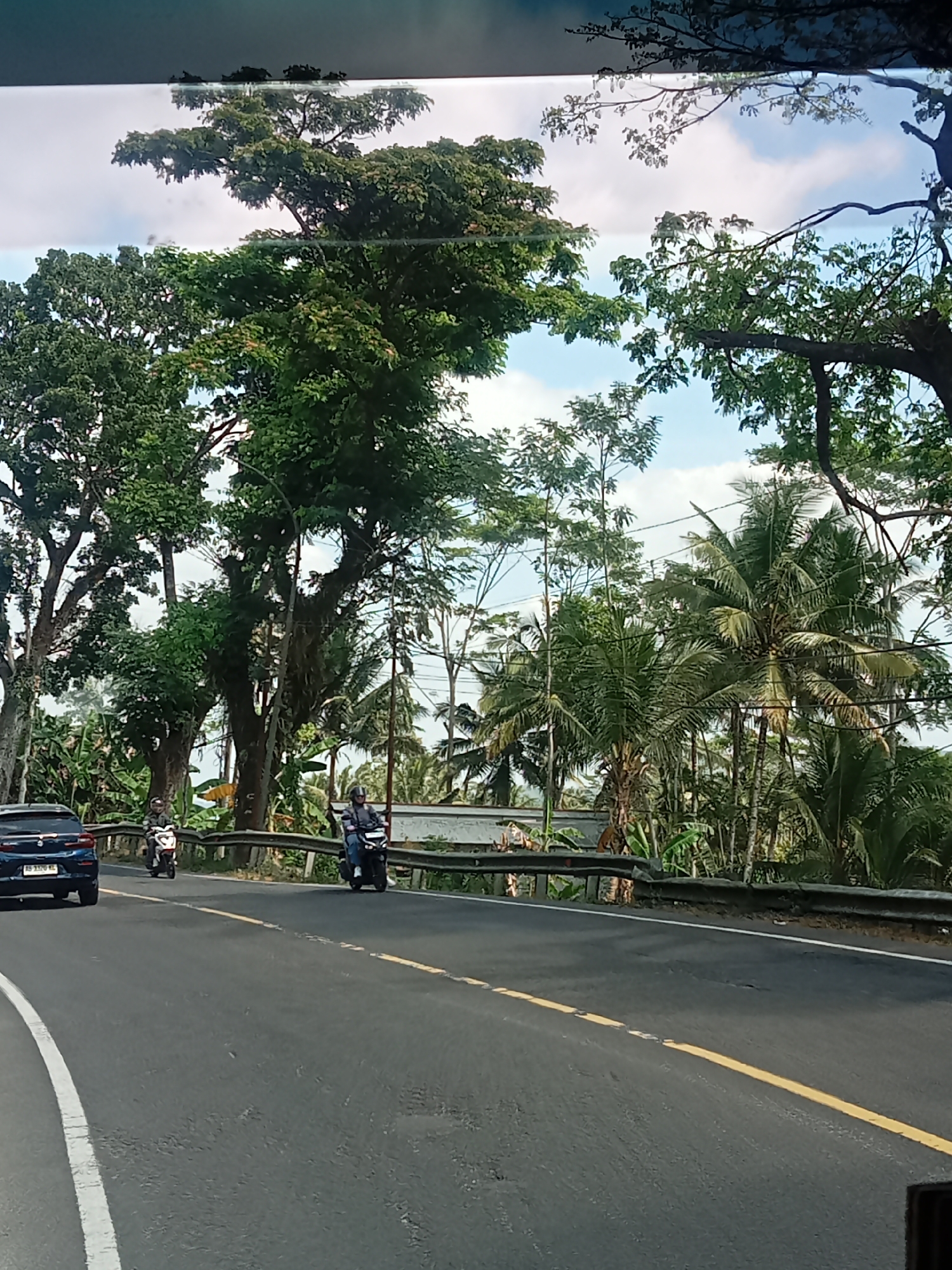 The image depicts a road scene in Selokromo, Indonesia. On the left side of the asphalt road, a dark blue car is visible. Further along the road, two individuals are riding motorcycles. The motorcycle on the left is white, and the one on the right is black. Both riders are wearing helmets. The road is bordered by a metal guardrail on the right side. Behind the guardrail, lush green foliage is abundant, with several large trees, palm trees, and banana plants. The sky above is partly cloudy with a bright blue hue. The overall environment suggests a tropical setting.