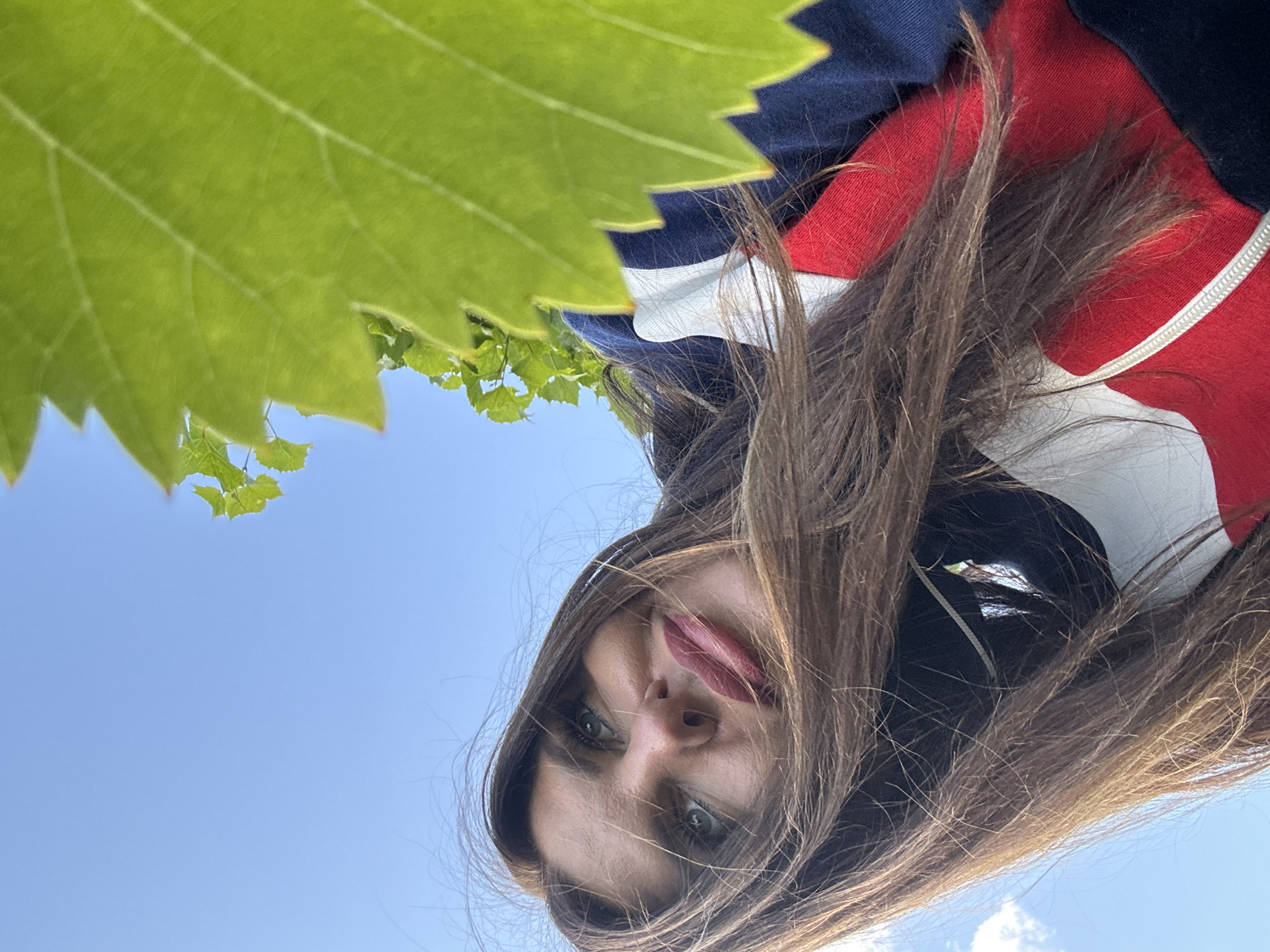 A low-angle, close-up, selfie-style photograph captures a woman from the chest up, outdoors, against a bright blue sky. She has long, brown hair that is partially obscuring her face, and she is wearing a dark blue, white, and red striped hooded jacket. A large, green leaf is prominently featured in the foreground, partially obscuring the woman's face and the sky. The woman is looking upwards and to her left, with her mouth slightly open. The setting appears to be a vineyard or an area with grapevines due to the presence of the grape leaf. The image does not contain any specific landmarks or contextual clues to pinpoint the location within Dubovaya, Russia, or any other city.