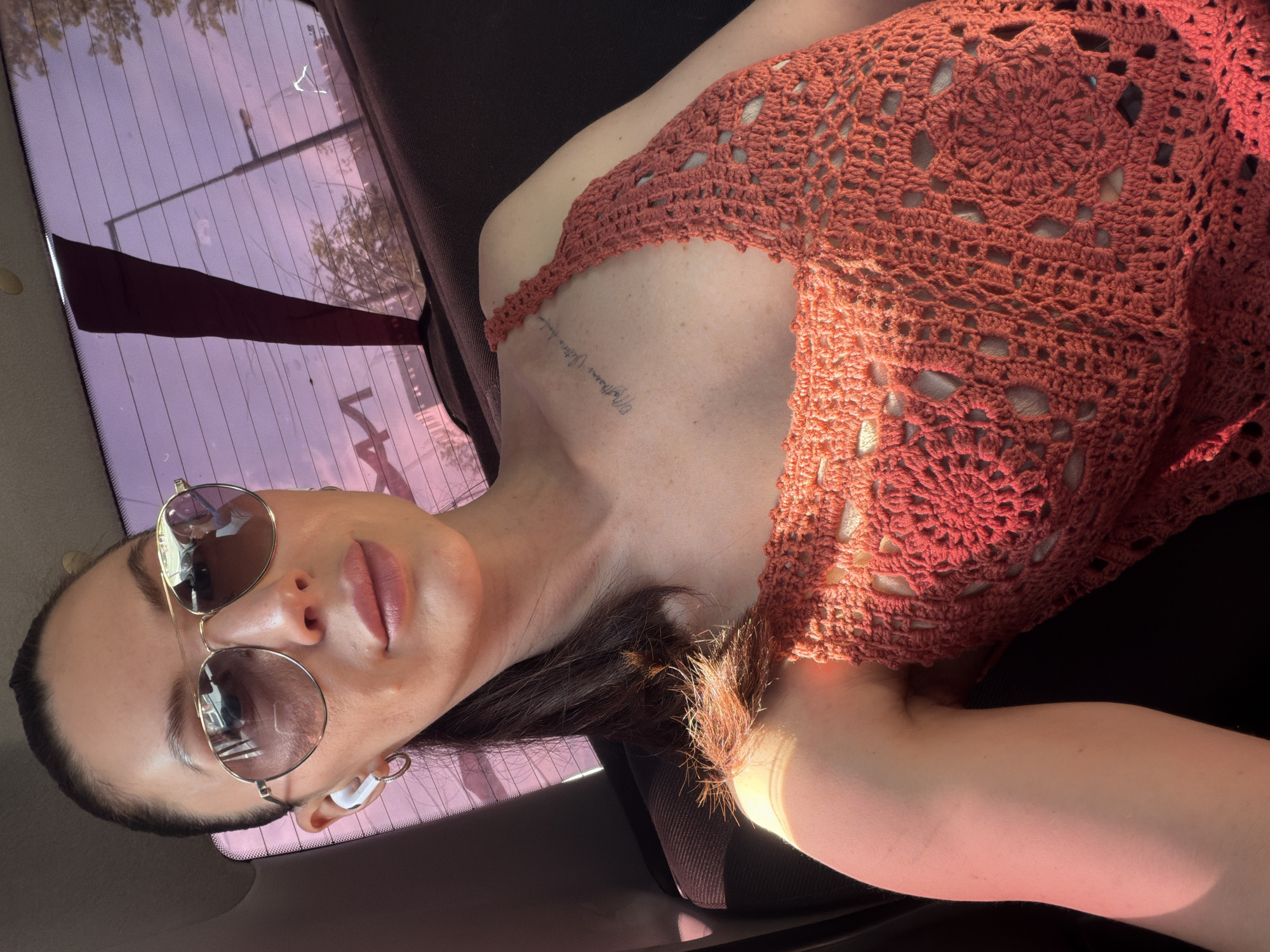 The image shows a woman in a car. She is wearing sunglasses and a reddish-orange crochet top. There's a tattoo visible on her chest. The car window behind her has a purplish tint, and glimpses of the outside world suggest an urban environment. The location is not clearly indicated as Perm, Russia.