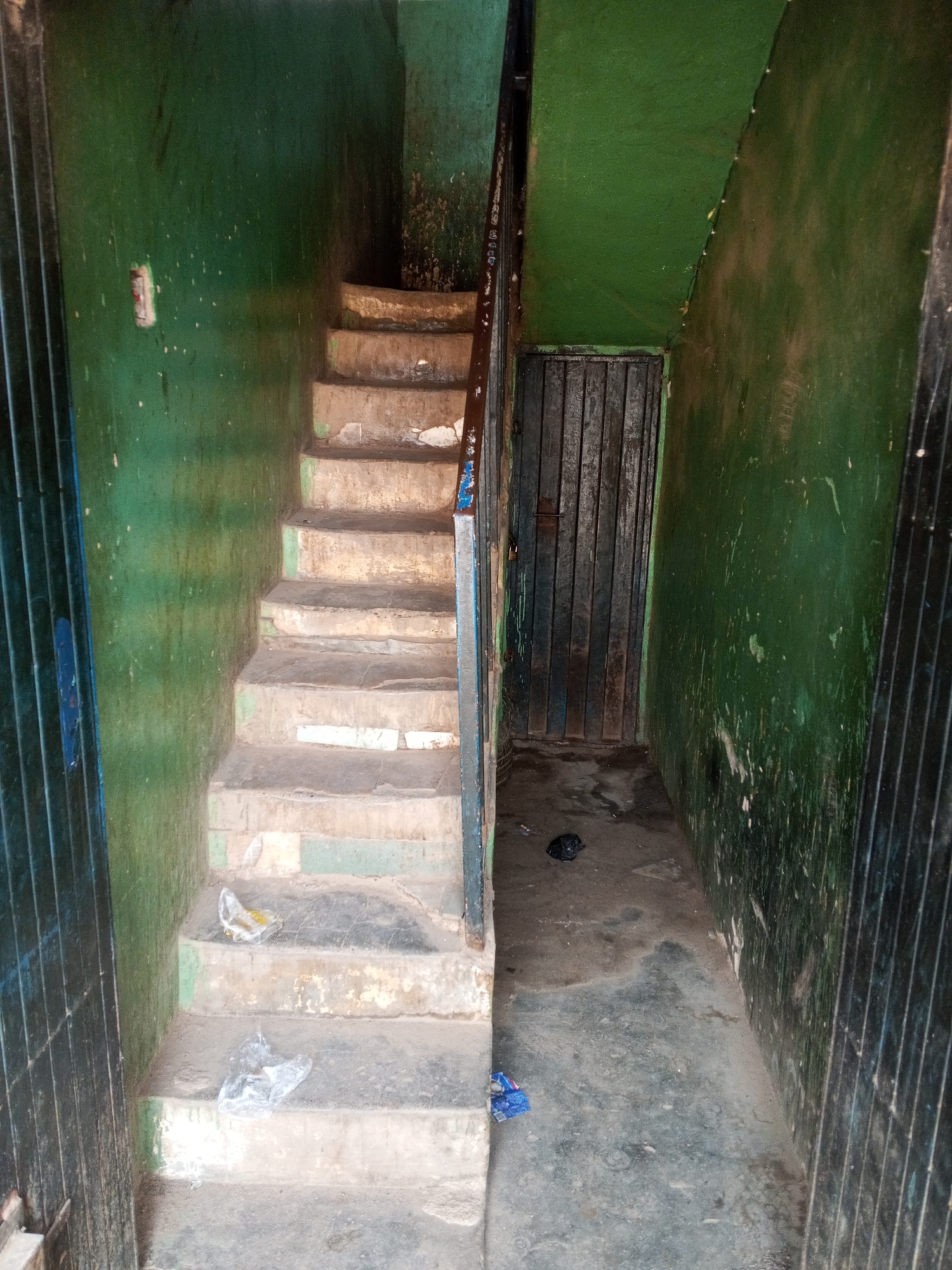 The image shows an interior staircase in a building in Gwong, Nigeria. The staircase is made of concrete and is in a state of disrepair. The walls are painted green but are stained and peeling. There is trash on the stairs and on the floor. A metal gate is visible on the left, and a metal door is on the right.