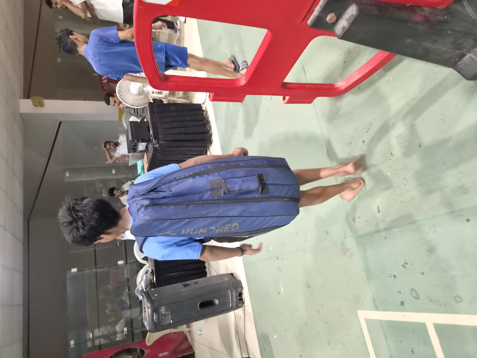 A young person, viewed from behind, walks across a green-colored court, carrying a large, blue sports bag over their right shoulder. The bag is emblazoned with the word "HUNDRED" in white lettering. The individual is wearing a blue t-shirt and dark shorts, and is barefoot. The background reveals a dimly lit indoor environment with several people present. To the left, a black speaker or equipment case stands near a table draped with a black cloth. In the middle ground, several individuals are visible, including one sitting near a fan. A bright red barrier is positioned in the foreground, partially obscuring the court. The scene appears to be in a sports facility, possibly a badminton or squash court, located within the city of Phagwara, India.