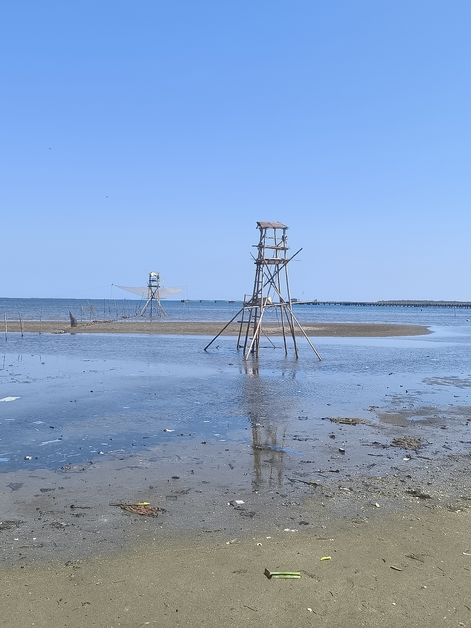 The image depicts a low-tide scene at a coastal area, likely in or near Barru, Indonesia, characterized by shallow water and exposed mudflats. In the foreground, the muddy shore is littered with debris, including plastic waste. The water is murky and reflects the sky and the structures present.
Dominating the mid-ground are two wooden observation or fishing structures. The structure on the right is a multi-tiered tower made of rough-hewn wooden poles, with a small, rustic roofed platform at the top. It is supported by angled legs planted in the shallow water and mud. A ladder is visible on its side. To the left, a similar, though slightly smaller and simpler, structure stands, also appearing to be made of wood and supported by poles. This structure has a large, triangular net or sail-like material attached to it, suggesting a possible fishing purpose.
In the background, a long, thin pier or causeway stretches across the water, lined with numerous vertical posts. The sea stretches to the horizon under a clear, pale blue sky, with a few distant ships or boats visible. The overall impression is one of a functional, utilitarian coastal environment, possibly used for fishing or observation, with the presence of pollution in the foreground indicating environmental concerns.