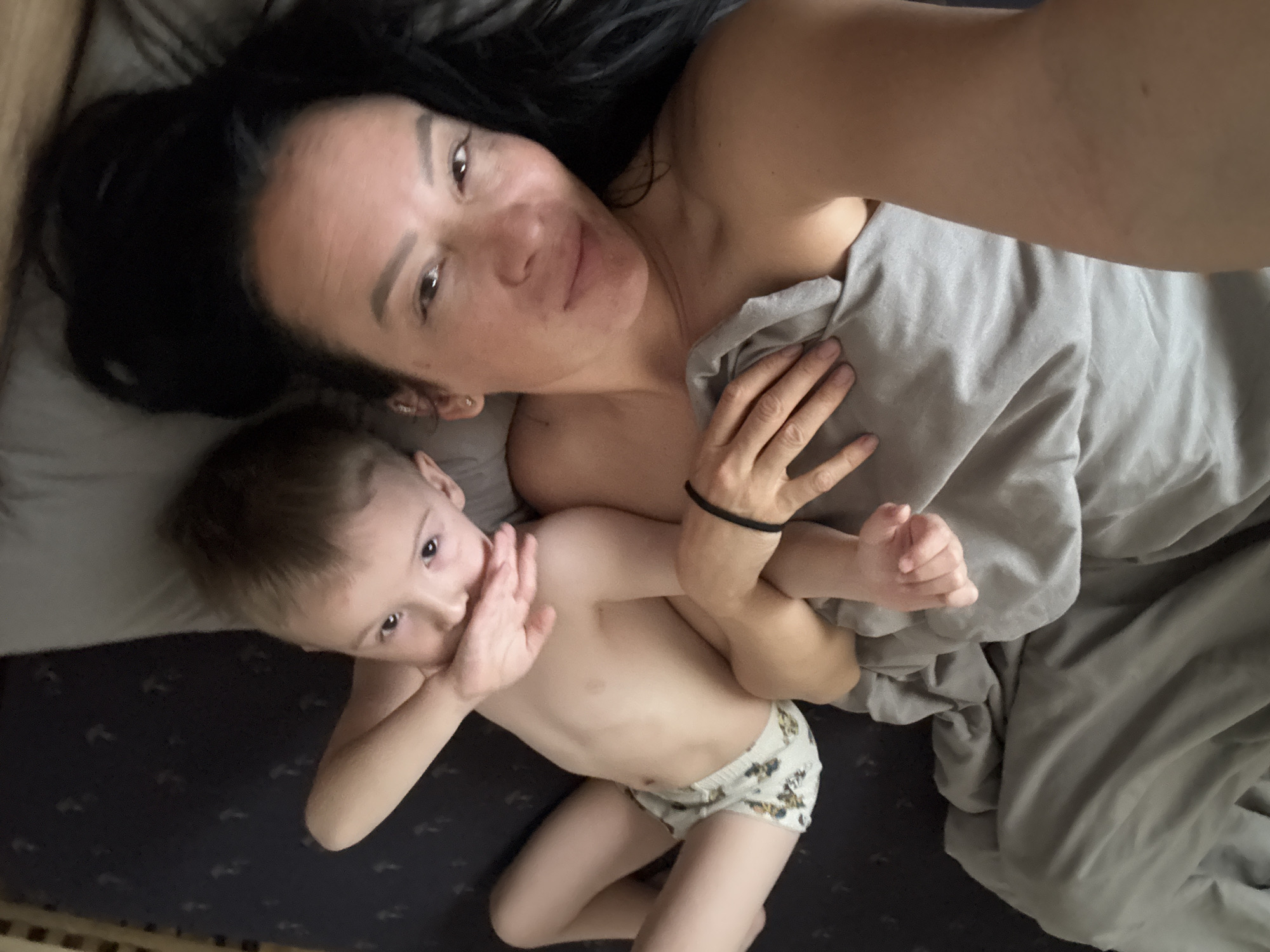 A woman with dark hair and a young child are lying in bed. The woman is facing the camera and smiling. The child is lying next to her, partially covered by a gray blanket. The location is a bed. The exact location in Chaikovsky, Russia is unknown.